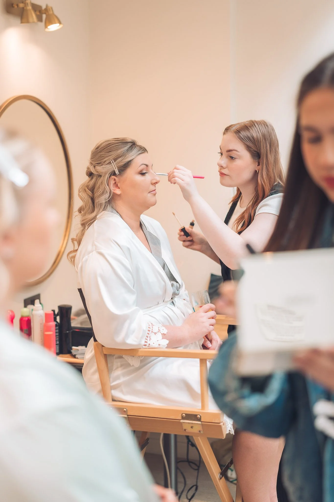 A woman with blonde hair getting her makeup done by a makeup artist in a room with beige walls and a mirror. The woman is seated in a wooden chair, wearing a white robe, holding a glass. The makeup artist is applying makeup around her nose, holding a