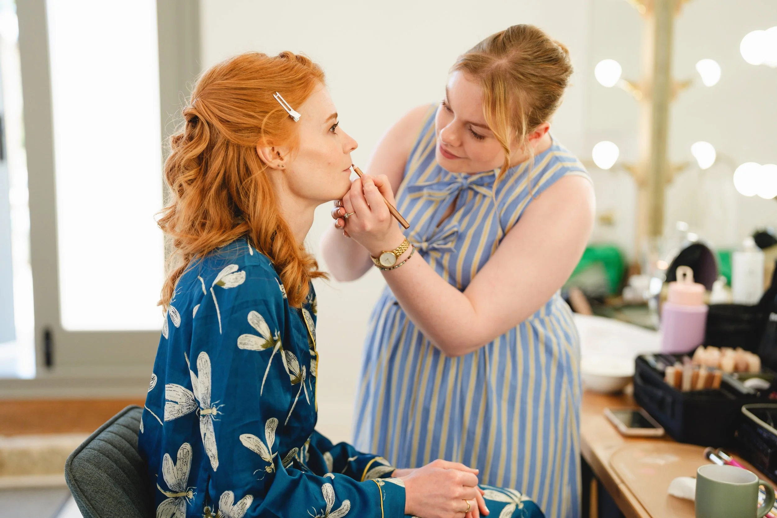 A makeup artist applying makeup to a woman with red hair and a blue floral robe, sitting in a makeup chair.