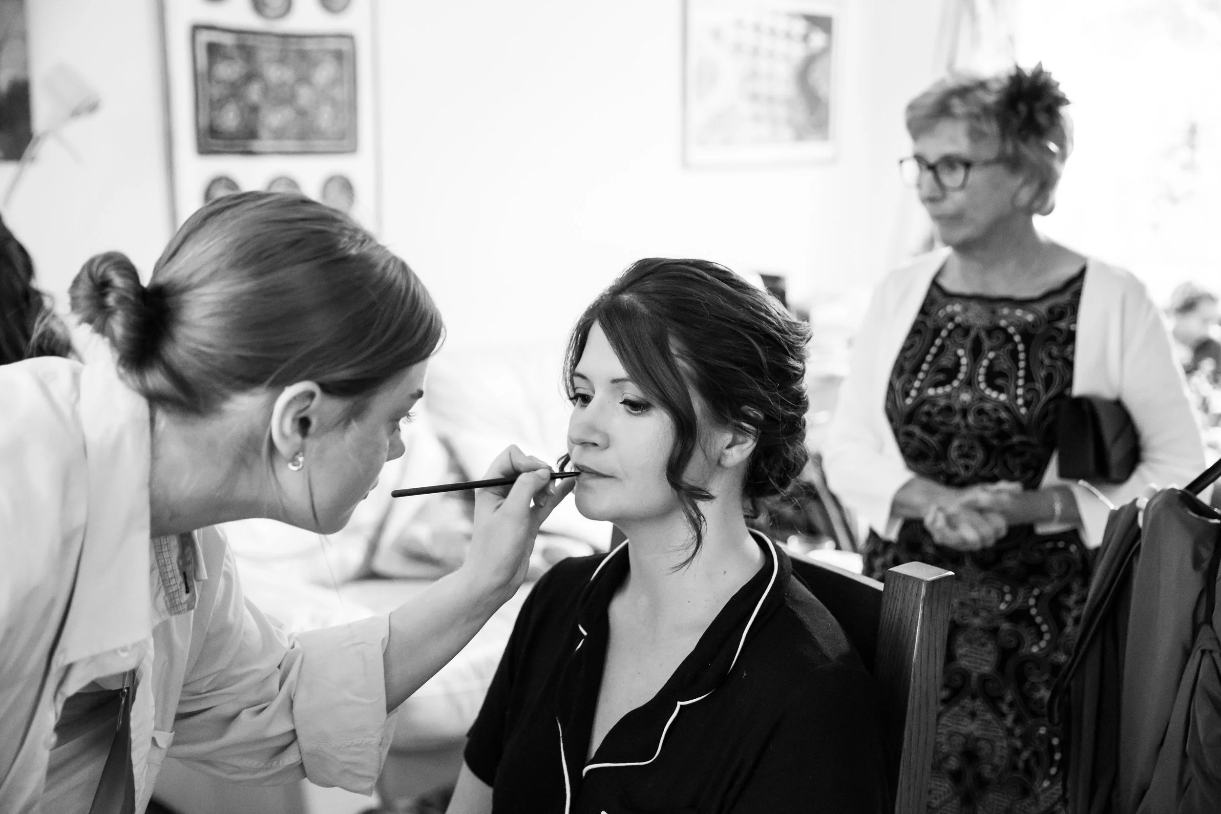 A woman getting her makeup done by a makeup artist, with another woman observing in the background.