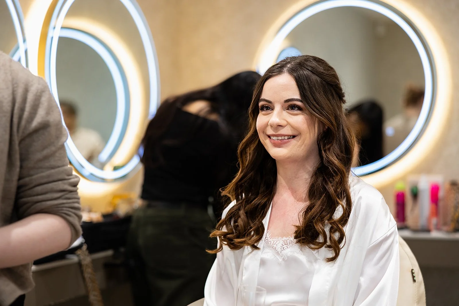 A young woman with long brown hair, smiling while sitting in a makeup salon, wearing a white satin robe. There are illuminated mirrors and various beauty products in the background.