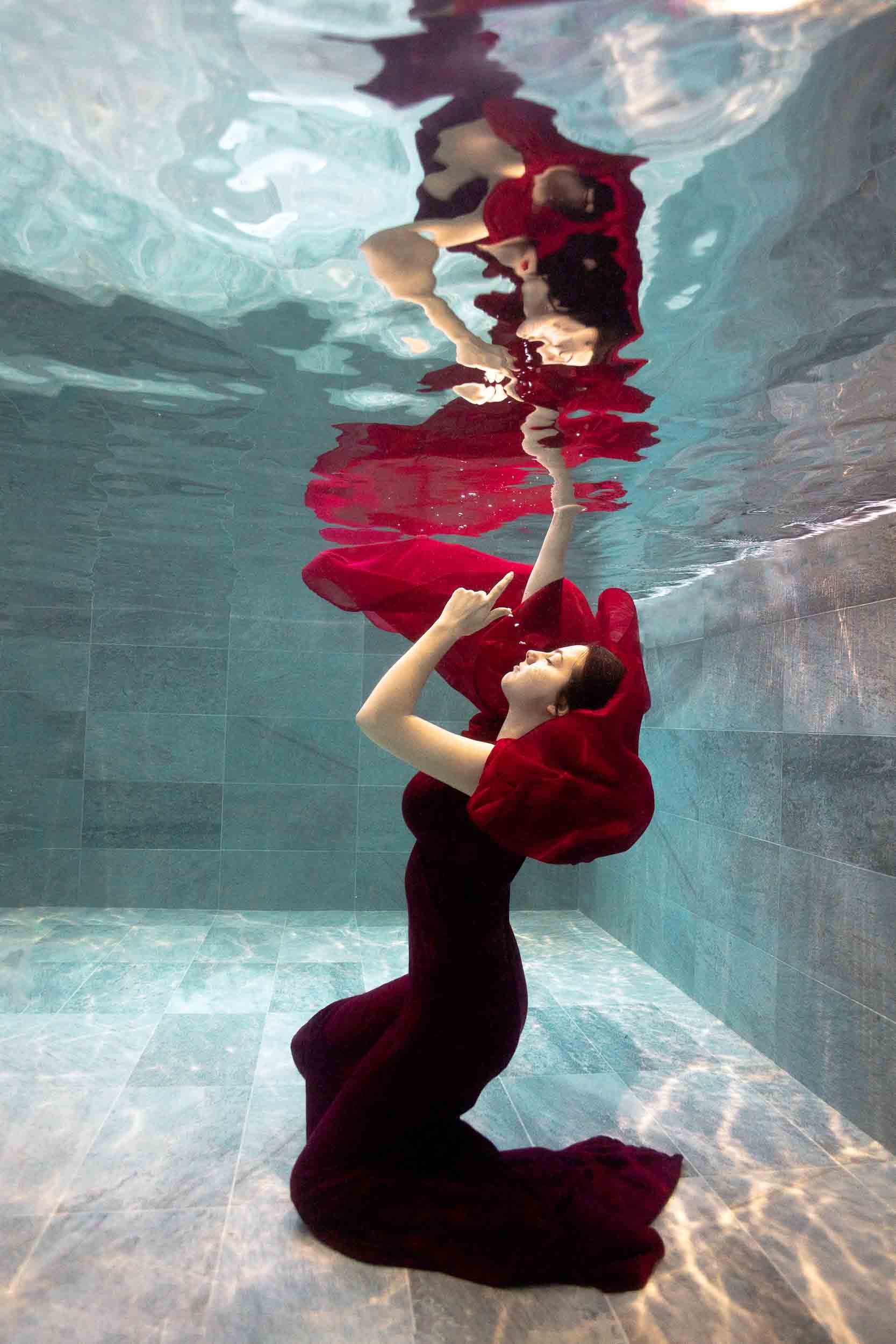 Une femme en robe rouge flotte sous l'eau, reflétée sur la surface. L'image est prise dans une piscine aux carreaux bleus.