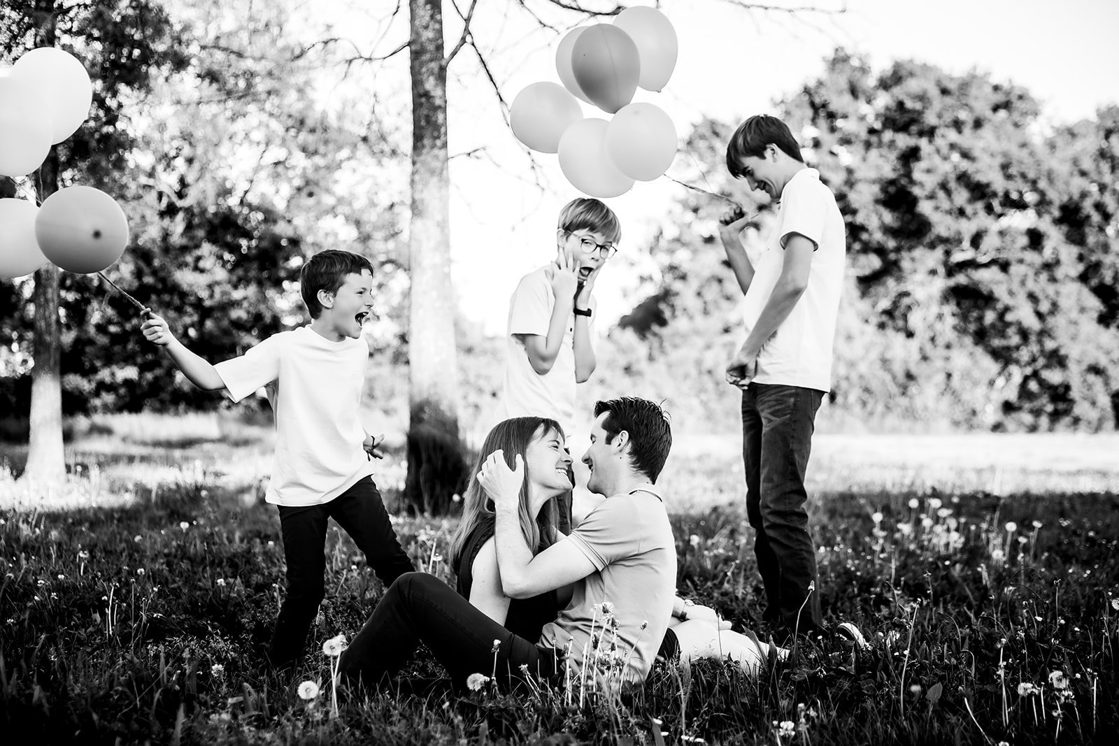 Couple souriant dans l'herbe avec leurs trois garcons les regardant en riant et tenant des ballons