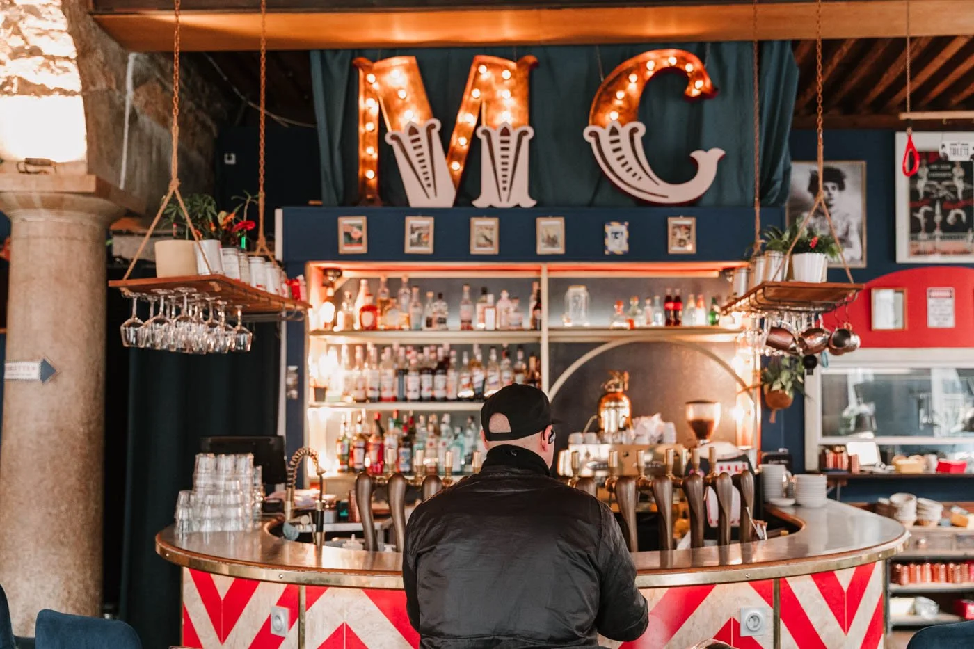 bar du restaurant le milord circus, en zinc et avec un motif blanc et rouge de cirque, un homme accoudé au bar de dos, et les initiales MC du milord circis en lettre de cirque lumineuse au dessus du comptoir