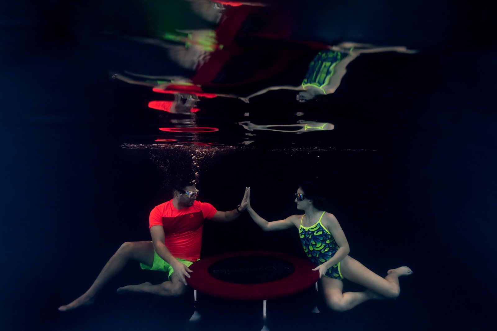 Deux personnes sous l'eau touchant leurs mains, portant des lunettes de natation et des vêtements colorés. Il y a un trampoline rouge entre elles. Leur reflet apparaît à la surface de l'eau.