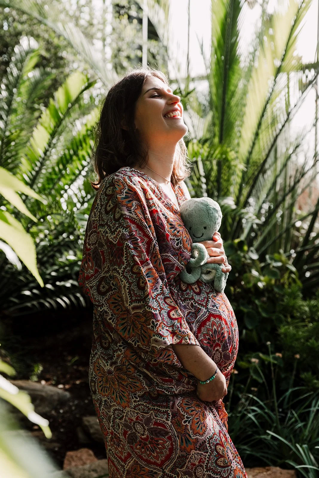 Femme enceinte riant dans une serre de plantes avec un doudou pieuvre bleu