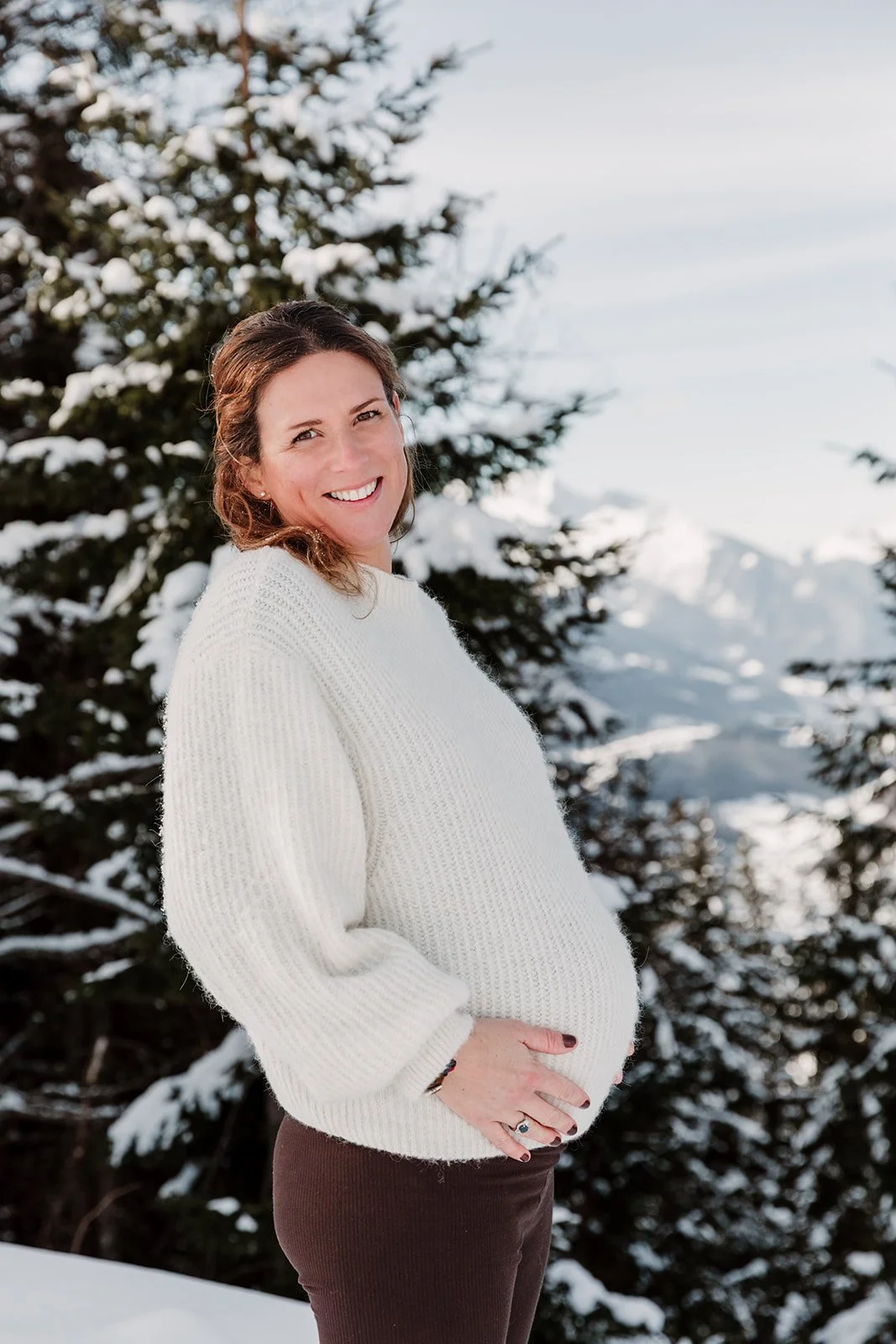 Femme enceinte souriante en pull blanc devant un paysage de montage et sapin enneigé
