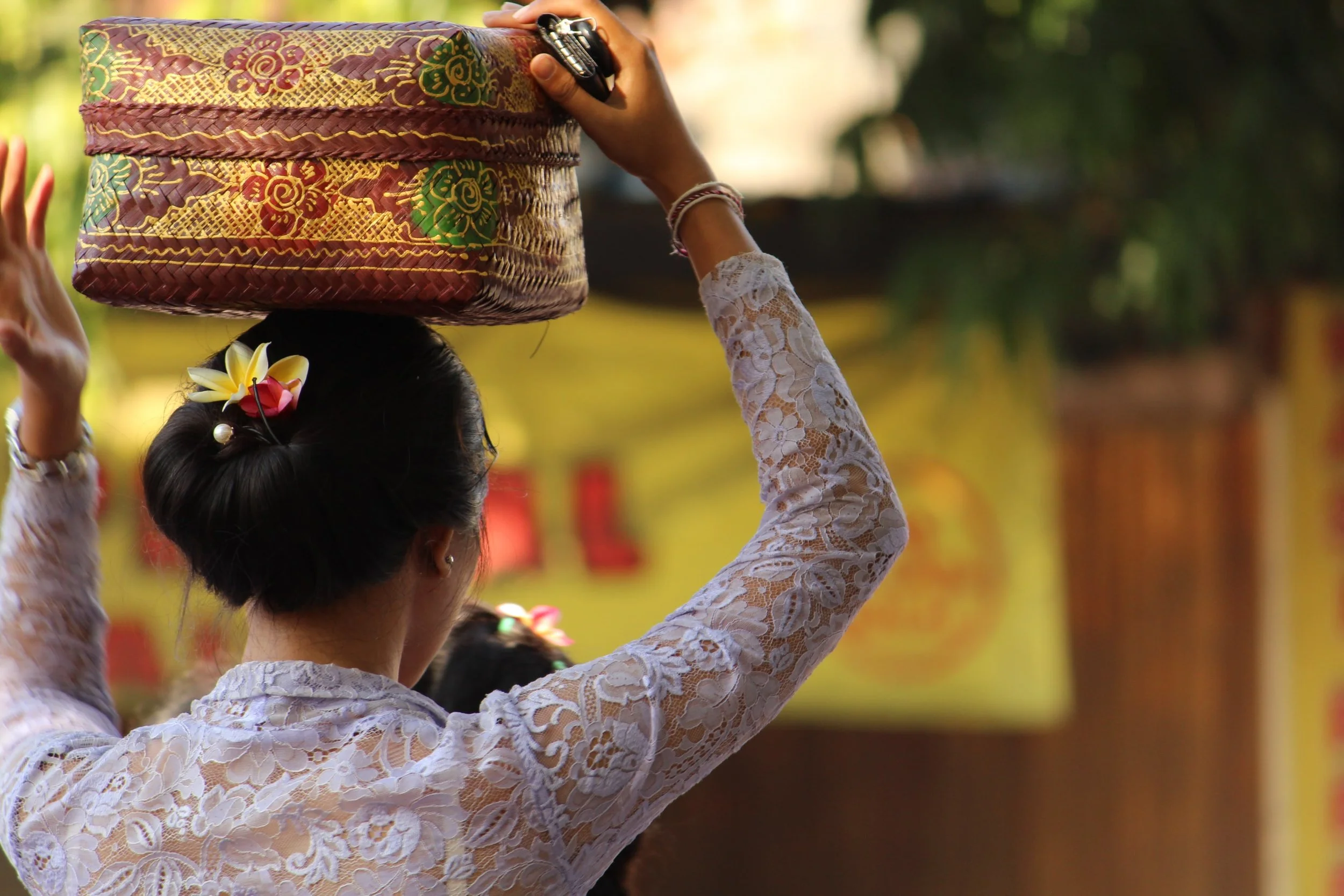 A woman dressed in a white lace traditional dress is balancing a colorful bag with intricate patterns on her head during a cultural event.