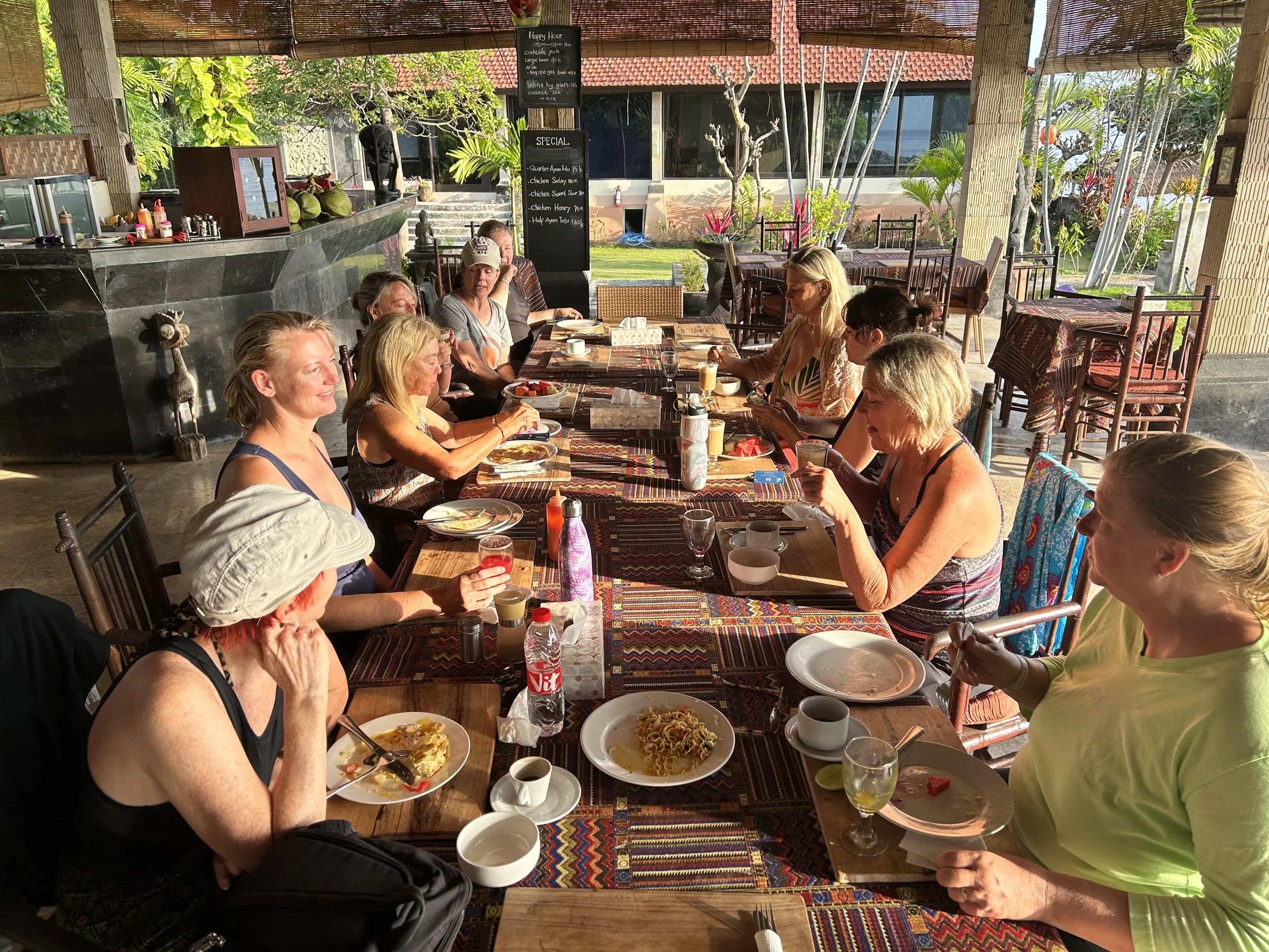 A group of people sitting around a long dining table outdoors, enjoying a meal. The table has various dishes, drinks, and condiments. The setting appears to be tropical, with lush greenery and a thatched-roof structure. The atmosphere is casual and lively.
