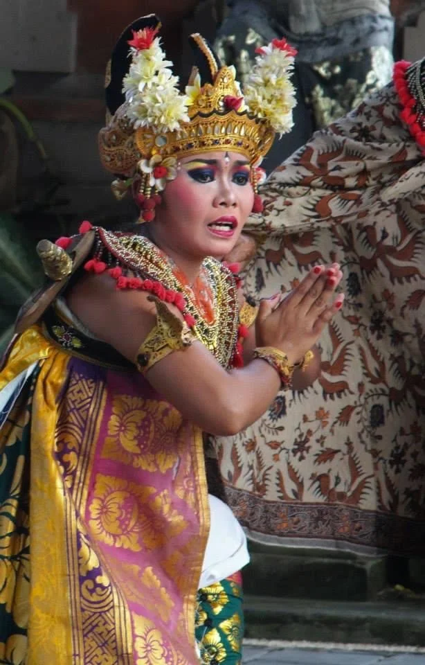 Young boy dressed as a traditional Hindu deity performing a prayer with hands pressed together, wearing an ornate headdress and colorful clothing with jewelry, during a cultural ceremony.