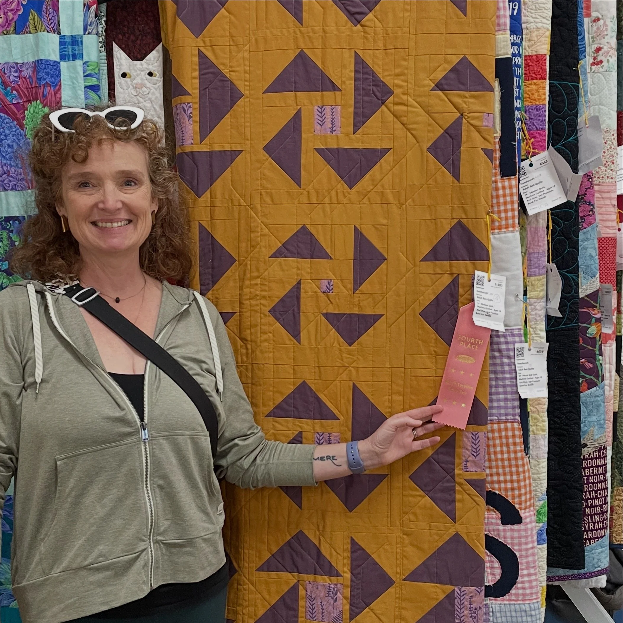Jenn, a middle-aged white woman with curly, greying red hair, stands proudly in front of a modern quilt with simple shapes in shades of purple on a gold background. Jenn is holding a peach-colored 4th place award ribbon attached to the quilt.