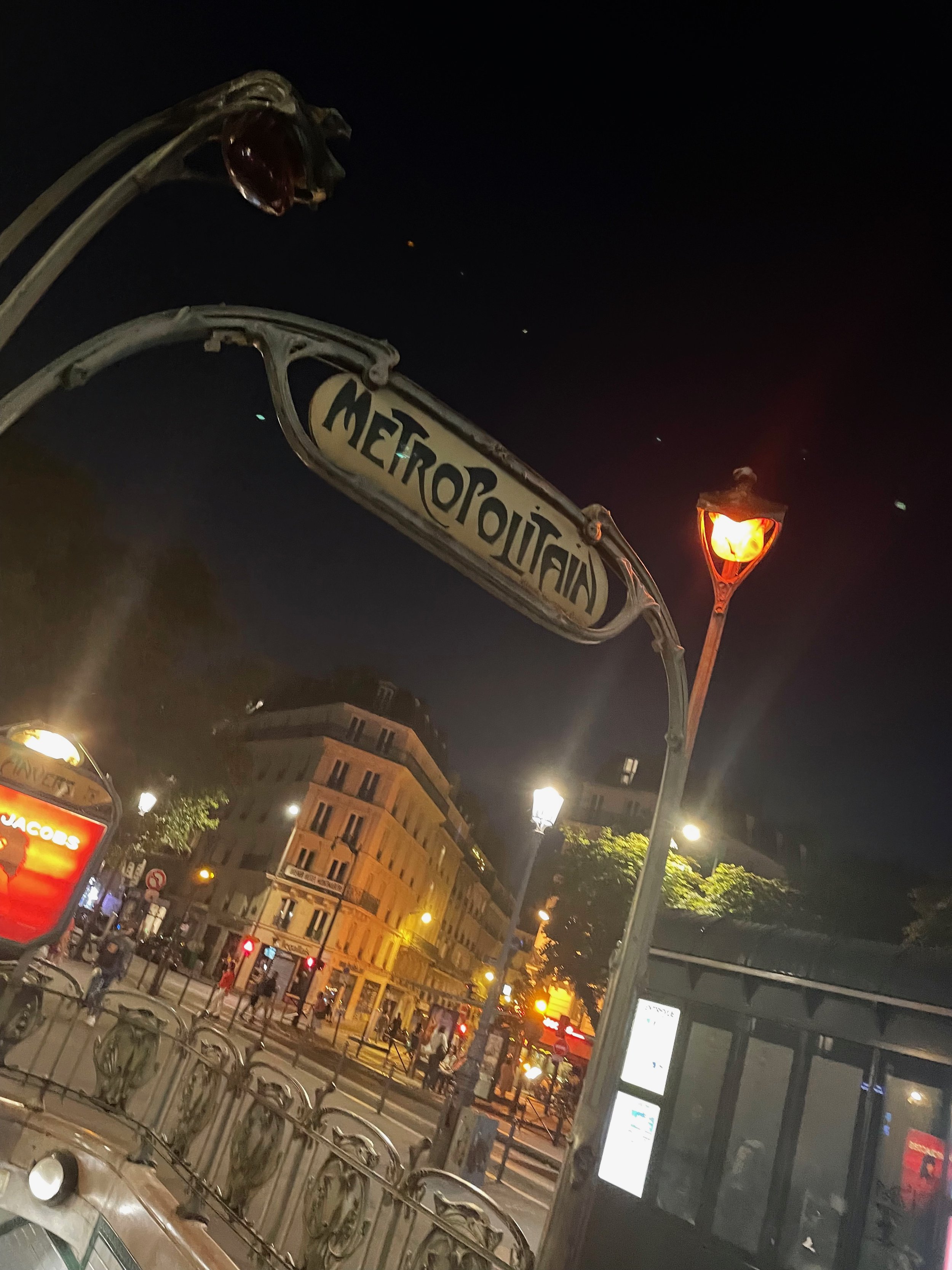  Image: An Art Nouveau style Metropolitain sign arches against a night sky, with buildings in the background. 
