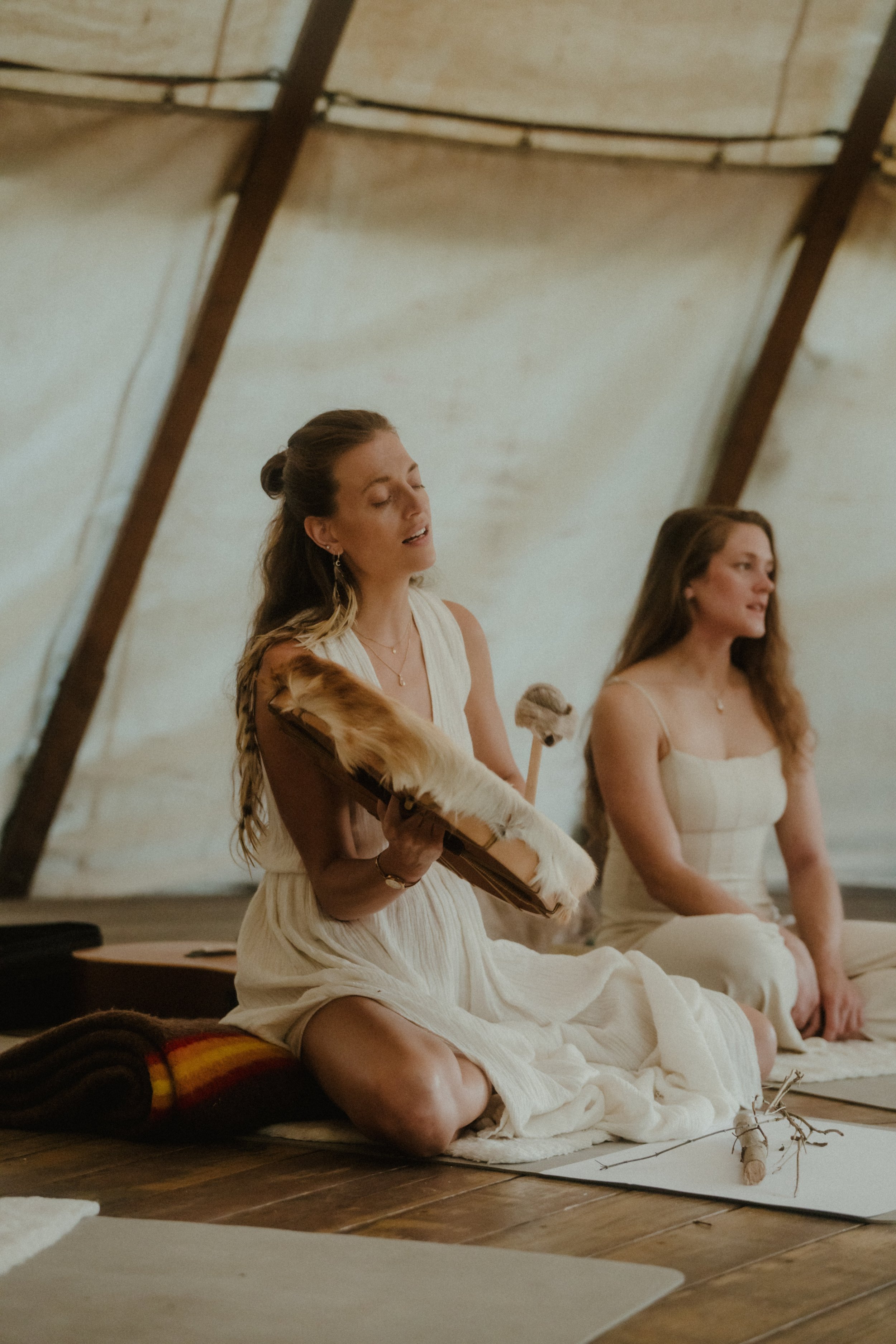 Two women sitting on the floor in a tent, with one playing a stringed instrument resembling a guitar, and the other with closed eyes, both dressed in cream-colored dresses, with blankets and branches on the floor, suggesting a spiritual or ceremonial gathering.