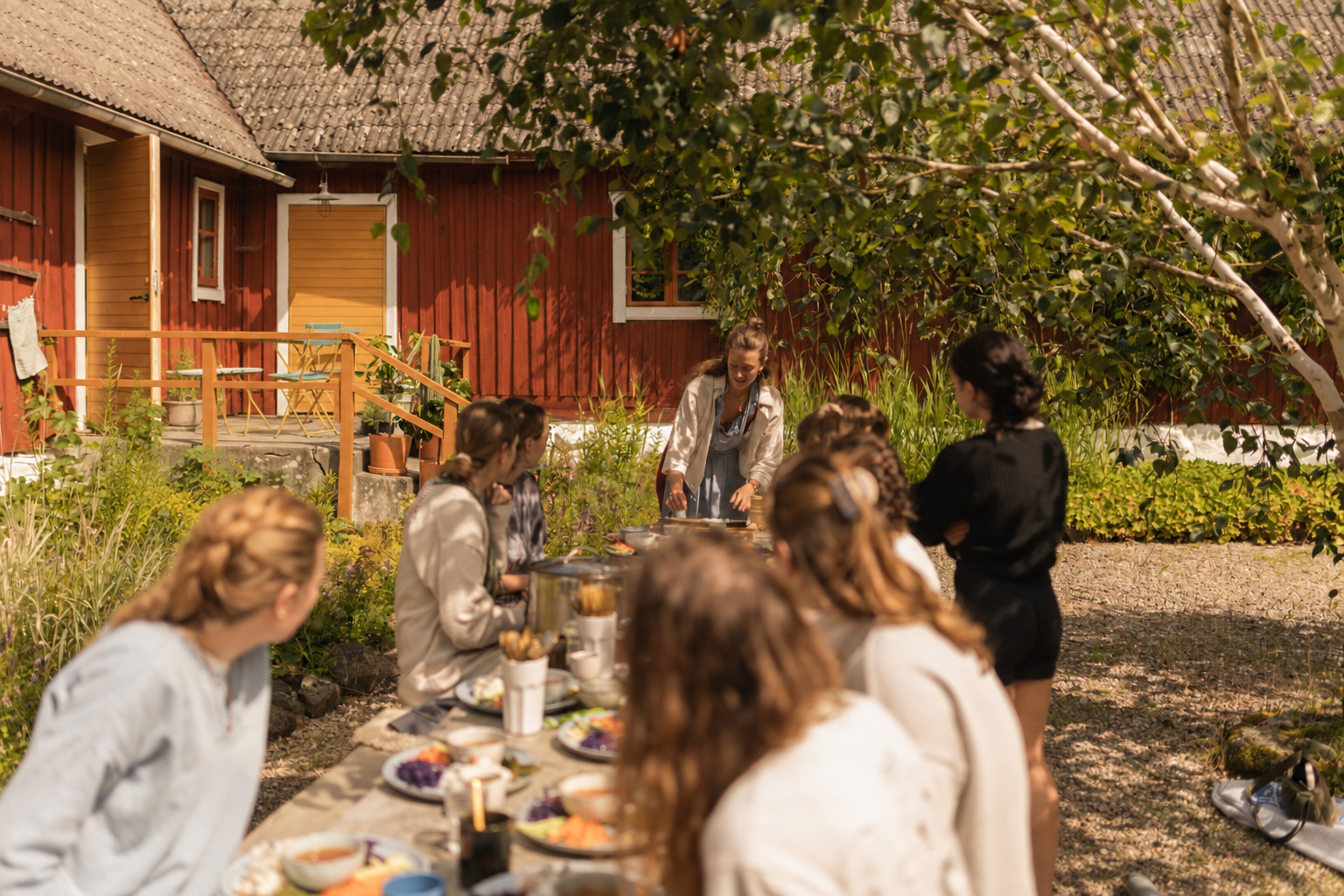 A group of young women gathered outdoors around a long table with food, participating in a social event or picnic near a red wooden house, with greenery and trees in the background.