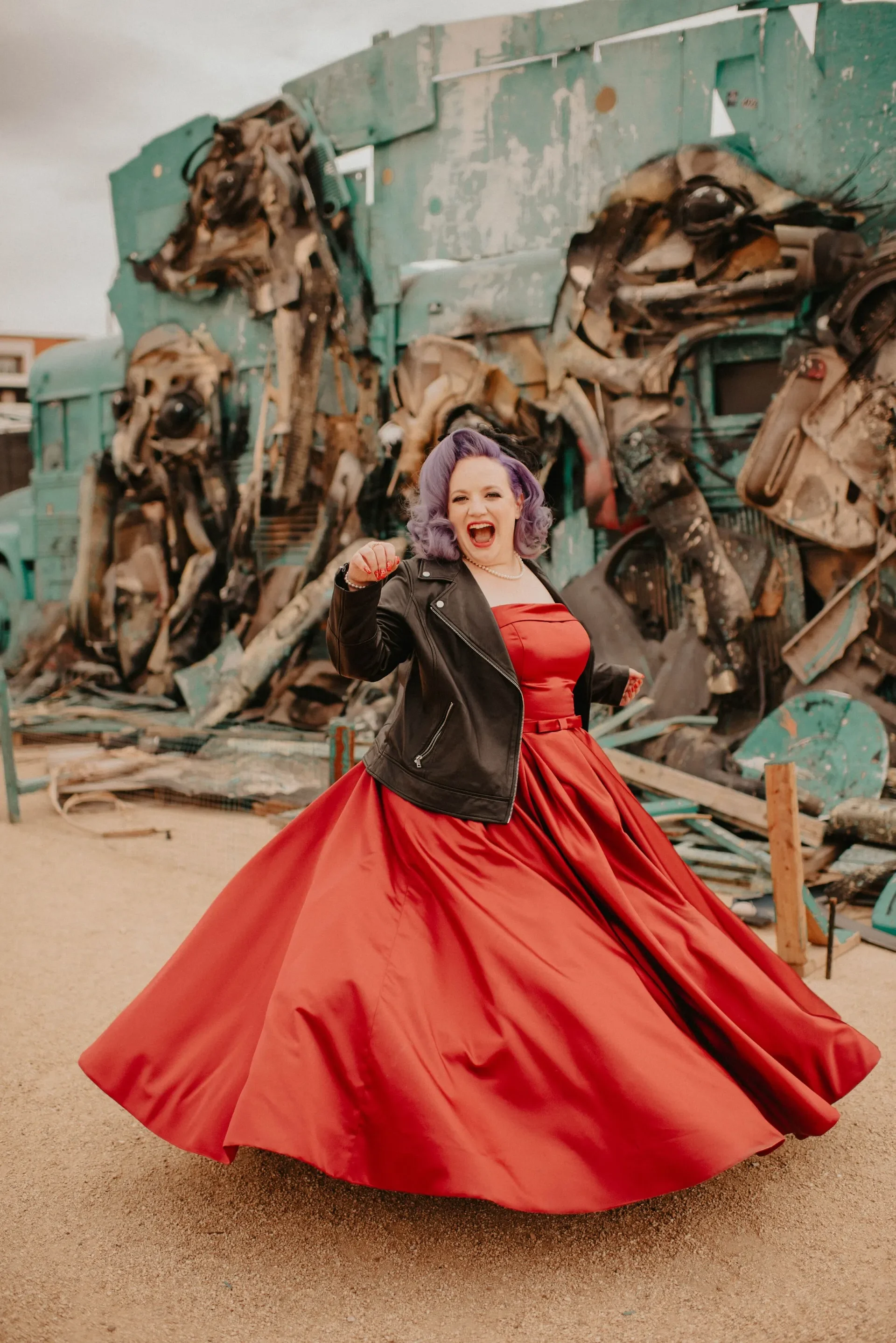 A woman with purple hair wearing a red ball gown and a black leather jacket, standing in front of a large, damaged, turquoise-colored vehicle, possibly a bus, with debris around.