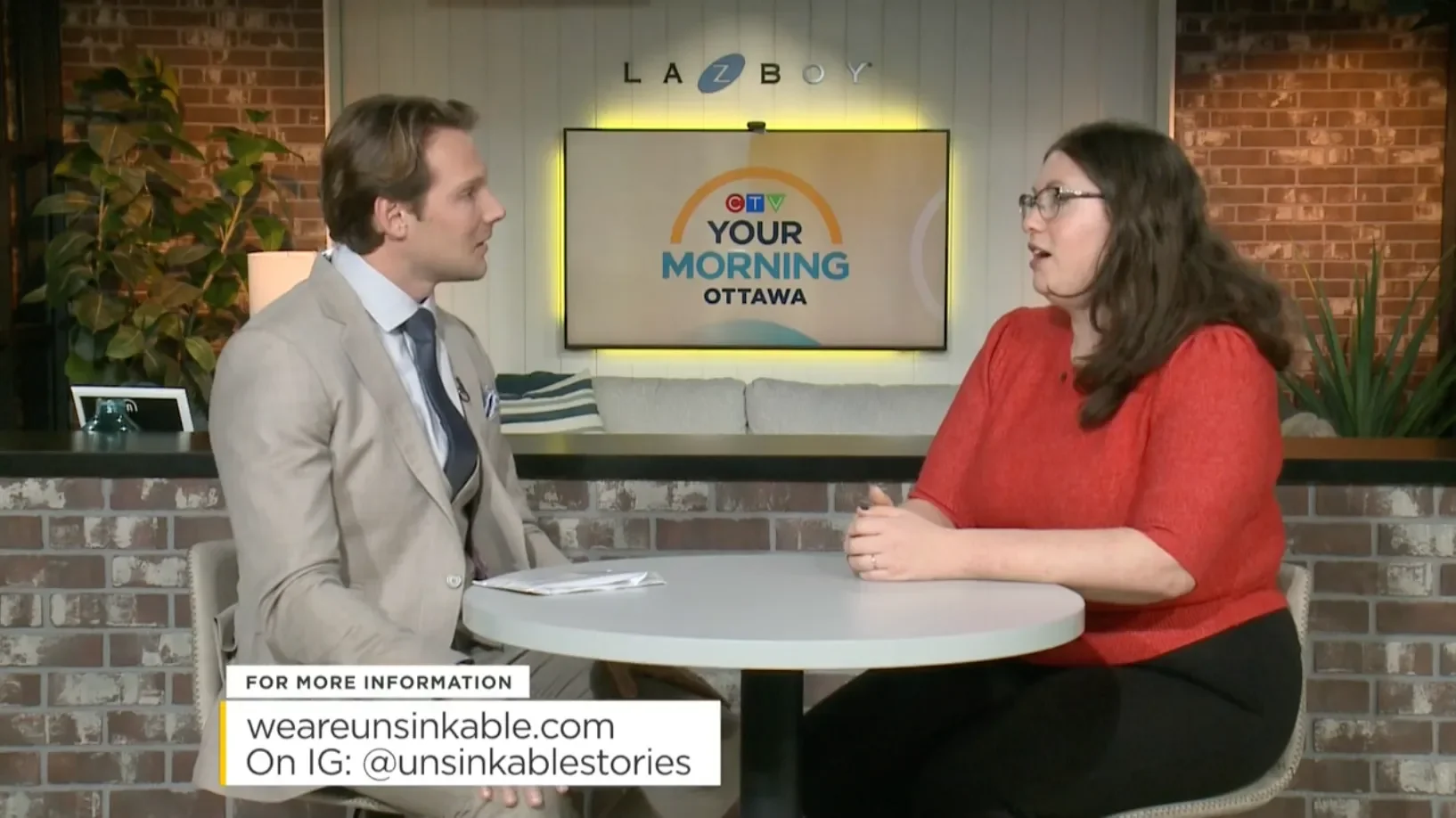 A man in a beige blazer talking to a woman, Hailey Hechtman during a morning television show in an urban studio with exposed brick walls, a TV monitor displaying the show's logo, Houseplants, and a table with a notebook in front of the man.