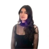 A woman with long dark hair wearing a black top with sheer polka-dot sleeves and a purple turtleneck underneath, standing against a white background.
