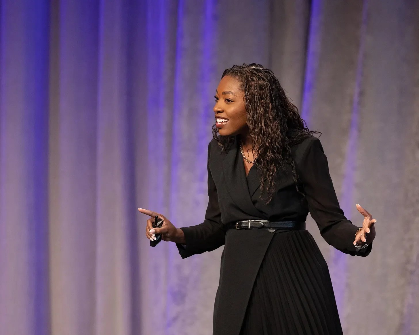 Woman in long black coat with belt smiling, giving a talk on a stage with a grey and purple backdrop
