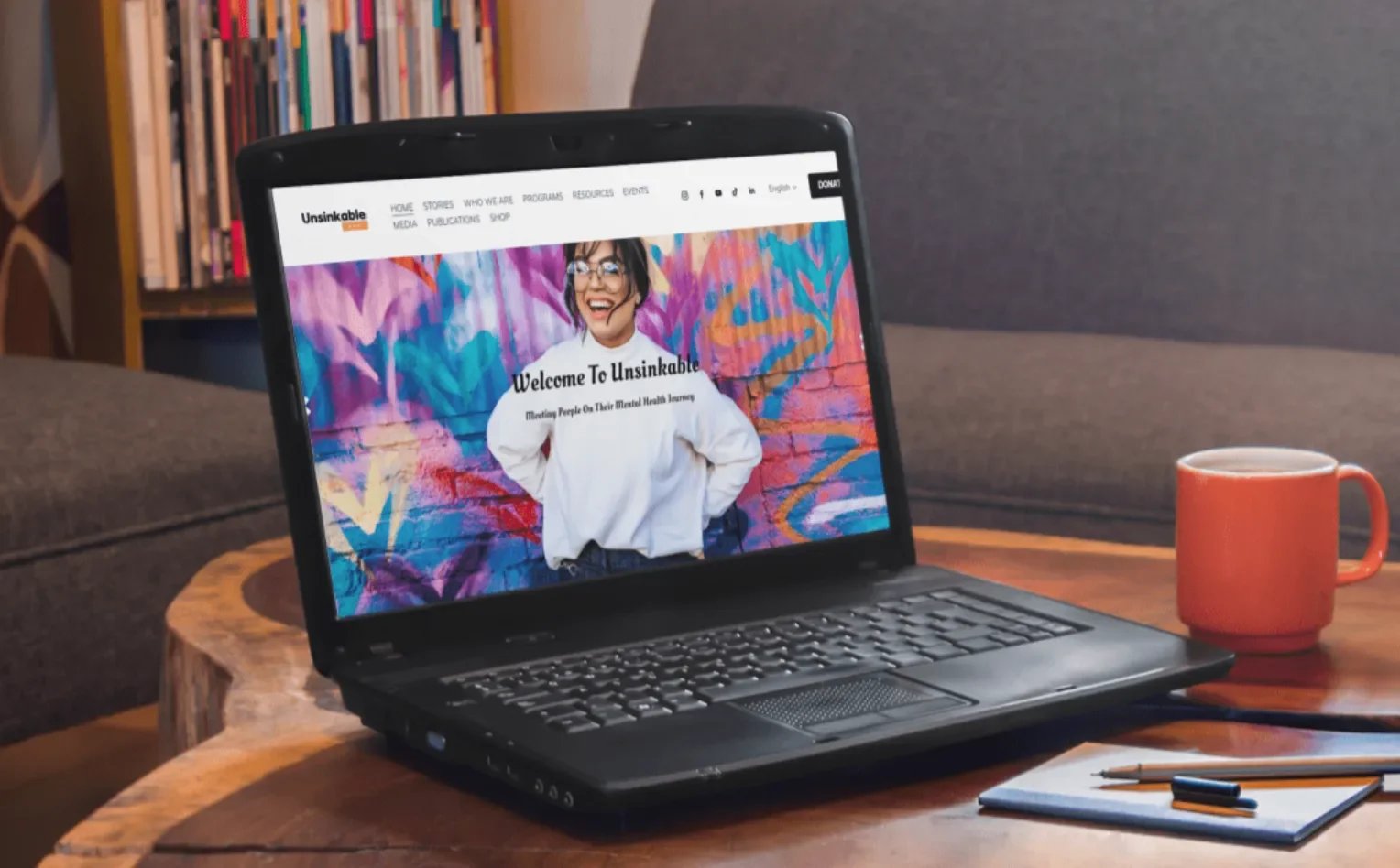 A laptop on a wooden table displaying a website with a smiling woman in front of a colorful graffiti wall. Next to the laptop is a red mug, and there are books, a notebook, and pens on the table.
