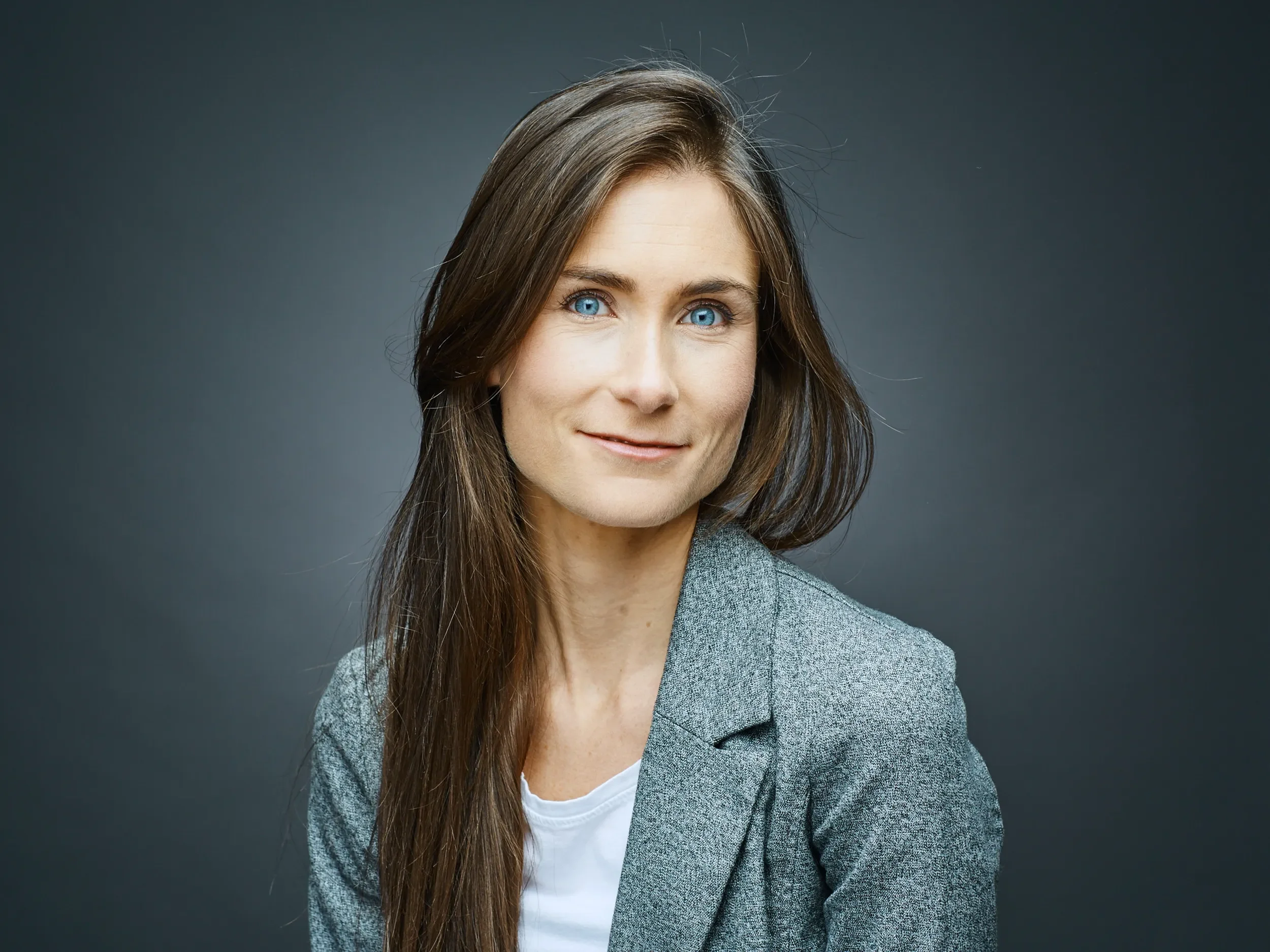 Portrait of a woman, Emma Sansfacon, with long brown hair, blue eyes, wearing a gray blazer and white top, standing against a dark gray background.