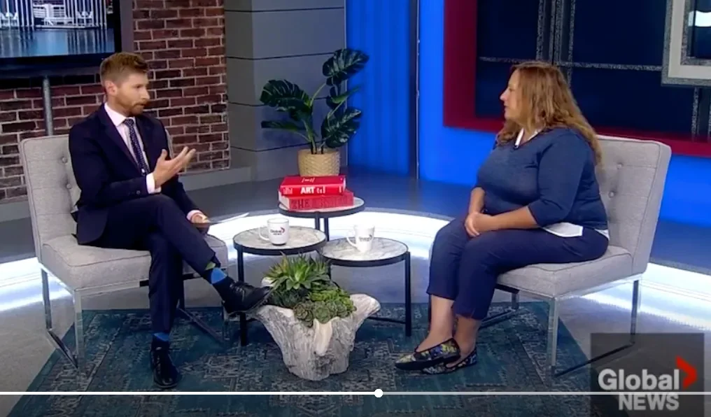 A man in a suit and a woman in a blue top are sitting on gray couches in a TV studio, engaged in conversation. There are two small round tables with mugs, a potted plant, and a stack of books between them. The background shows a brick wall and a window.
