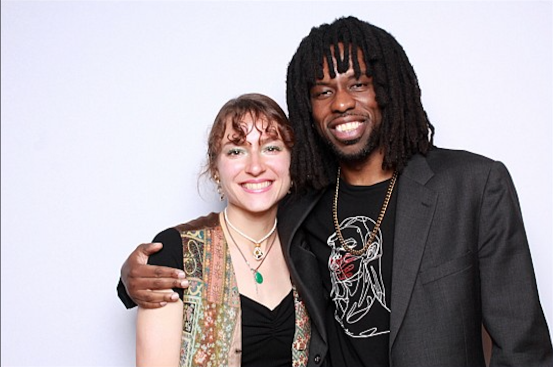 A man with dark skin and dreadlocked hair, wearing a graphic tee and a dark blazer, and a woman with curly hair and a patterned vest, smiling into the camera against a light background.