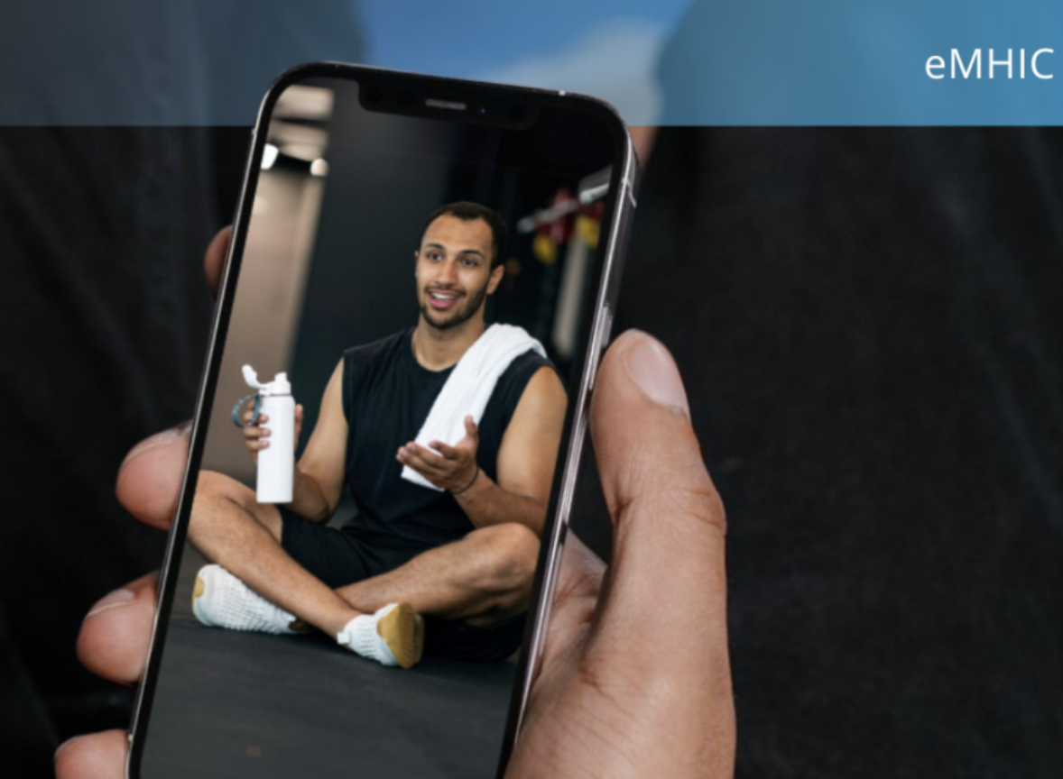 Person holding a smartphone taking a selfie with a man sitting cross-legged holding a water bottle in a gym or fitness studio.