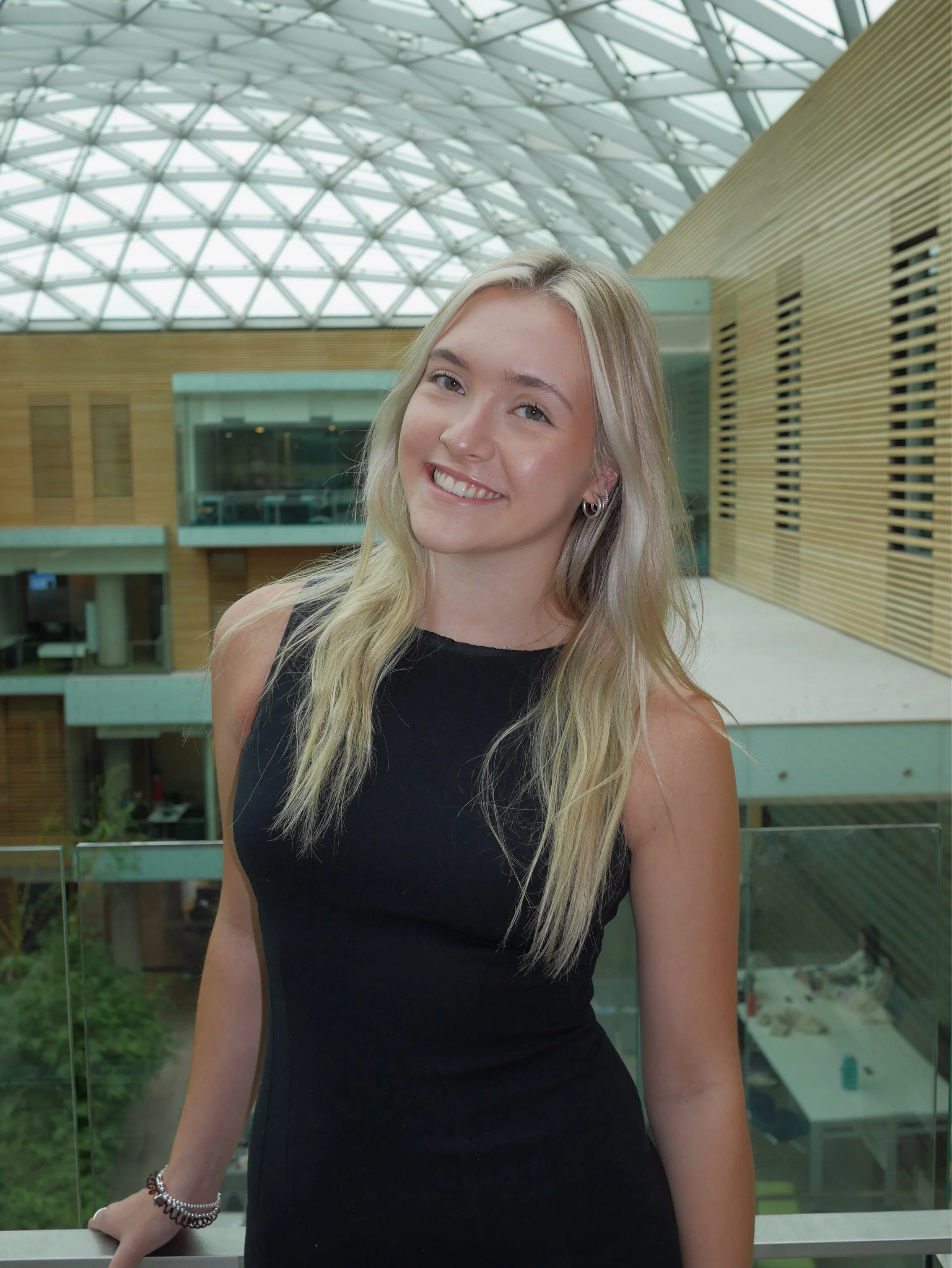 A young woman with long blonde hair wearing a sleeveless black dress, standing inside a modern building with a glass ceiling and wooden wall panels, smiling at the camera.