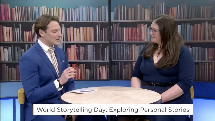 A man and a woman sit at a small round table in a studio with bookshelves background. The man is wearing a blue suit with a light-colored striped tie, and the woman is in a dark blue dress. The caption reads: 'World Storytelling Day: Exploring Personal Stories.'