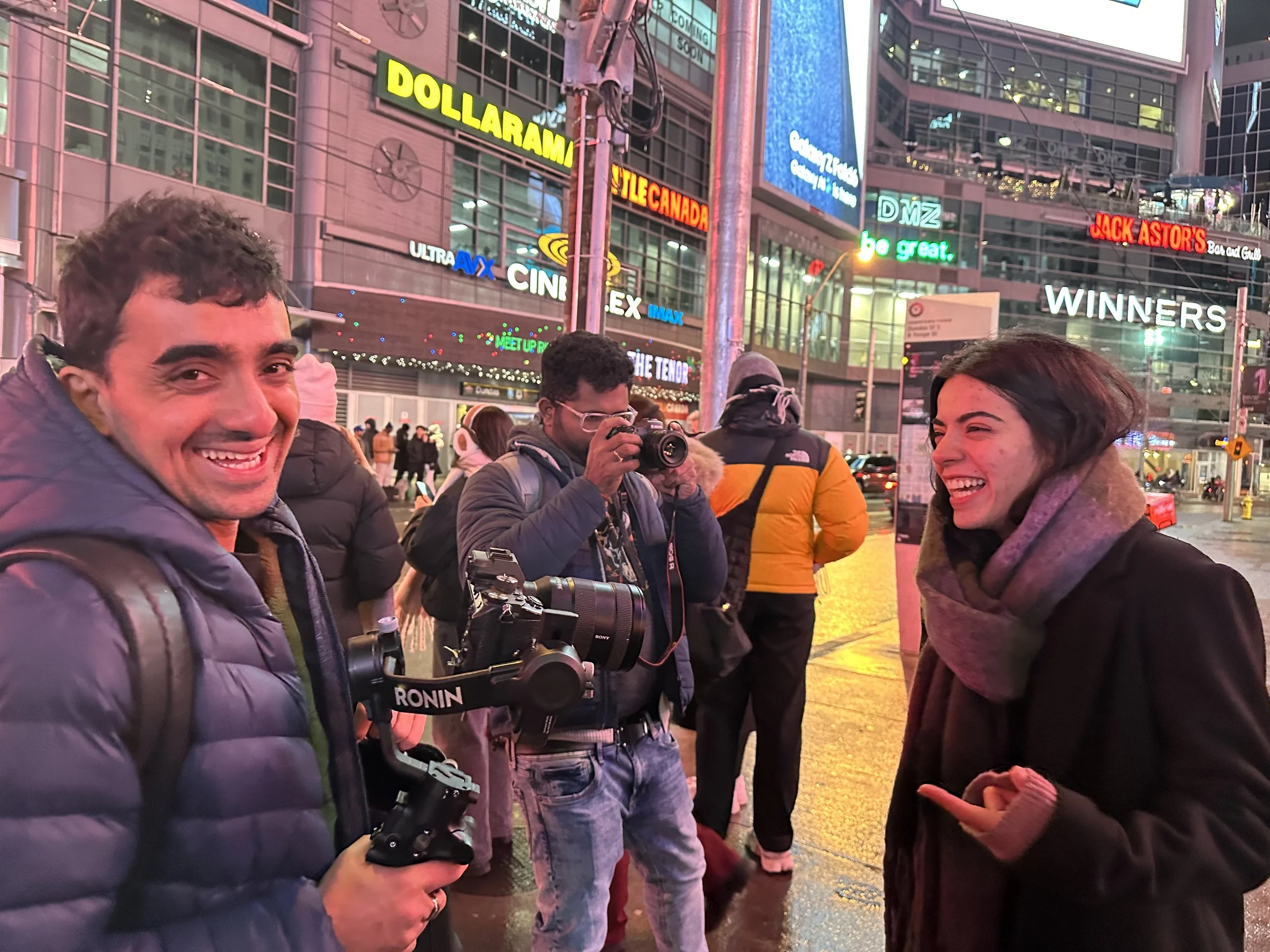 Group of young adults standing together on a busy city street at night, with colorful neon signs and tall buildings in the background. One person is taking a photo, while others are smiling and talking.