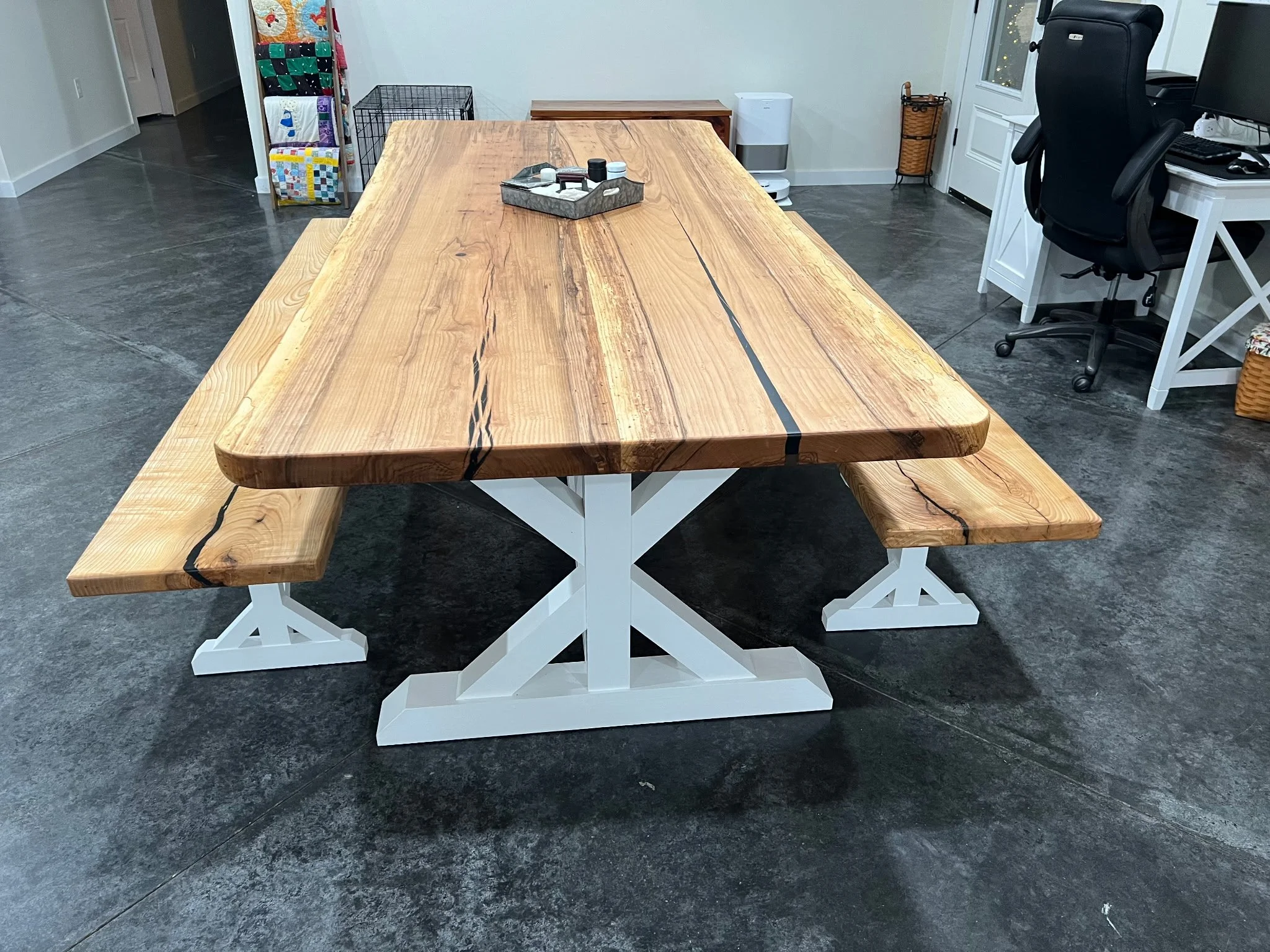 A large wooden dining table with a natural finish and white painted trestle base, surrounded by a matching wooden bench, situated on a dark concrete floor in a modern interior space.