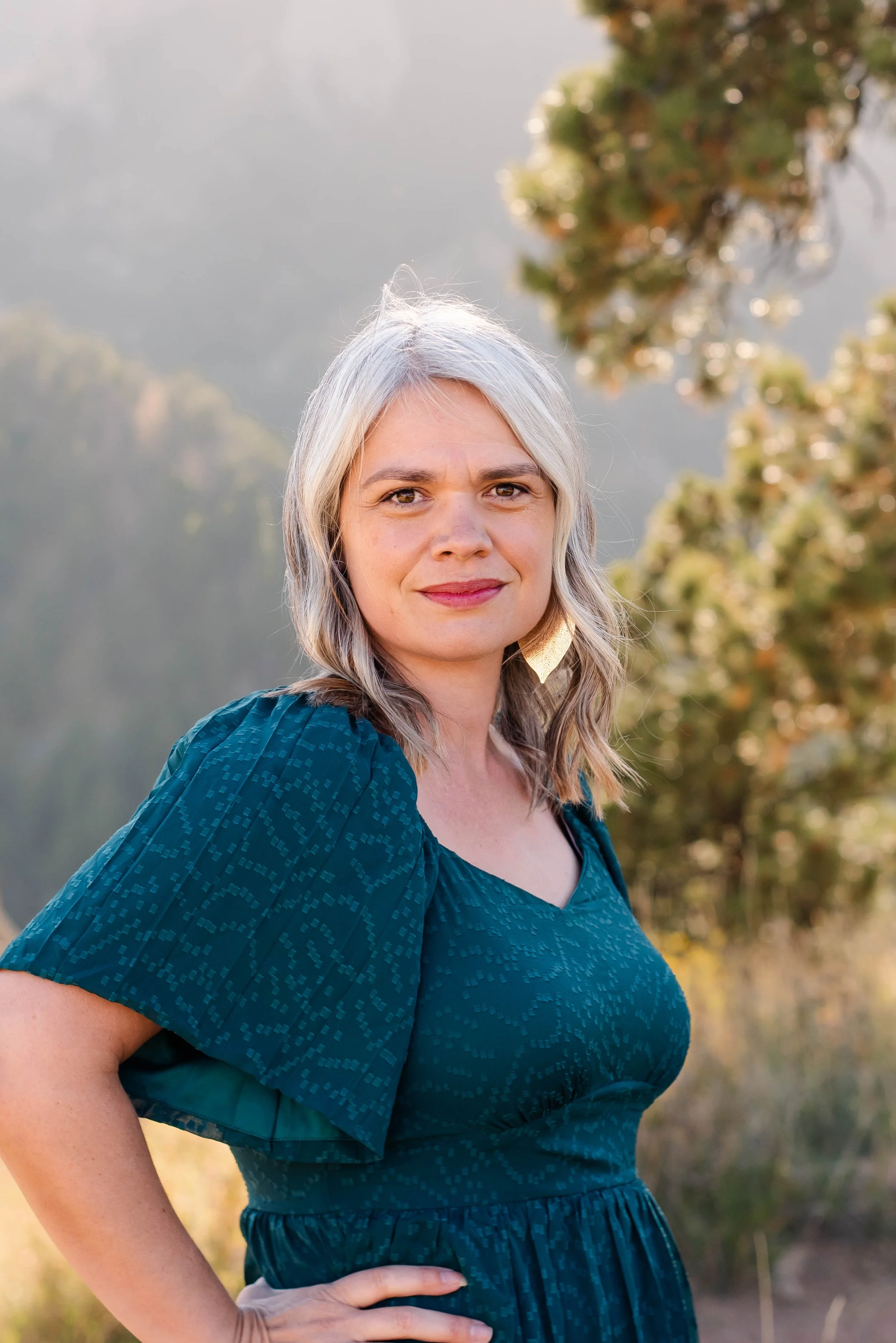 A woman with shoulder-length gray hair standing outdoors with her hand on her hip, wearing a teal dress with puffed sleeves and a subtle pattern, in front of a natural background with trees and mountains.