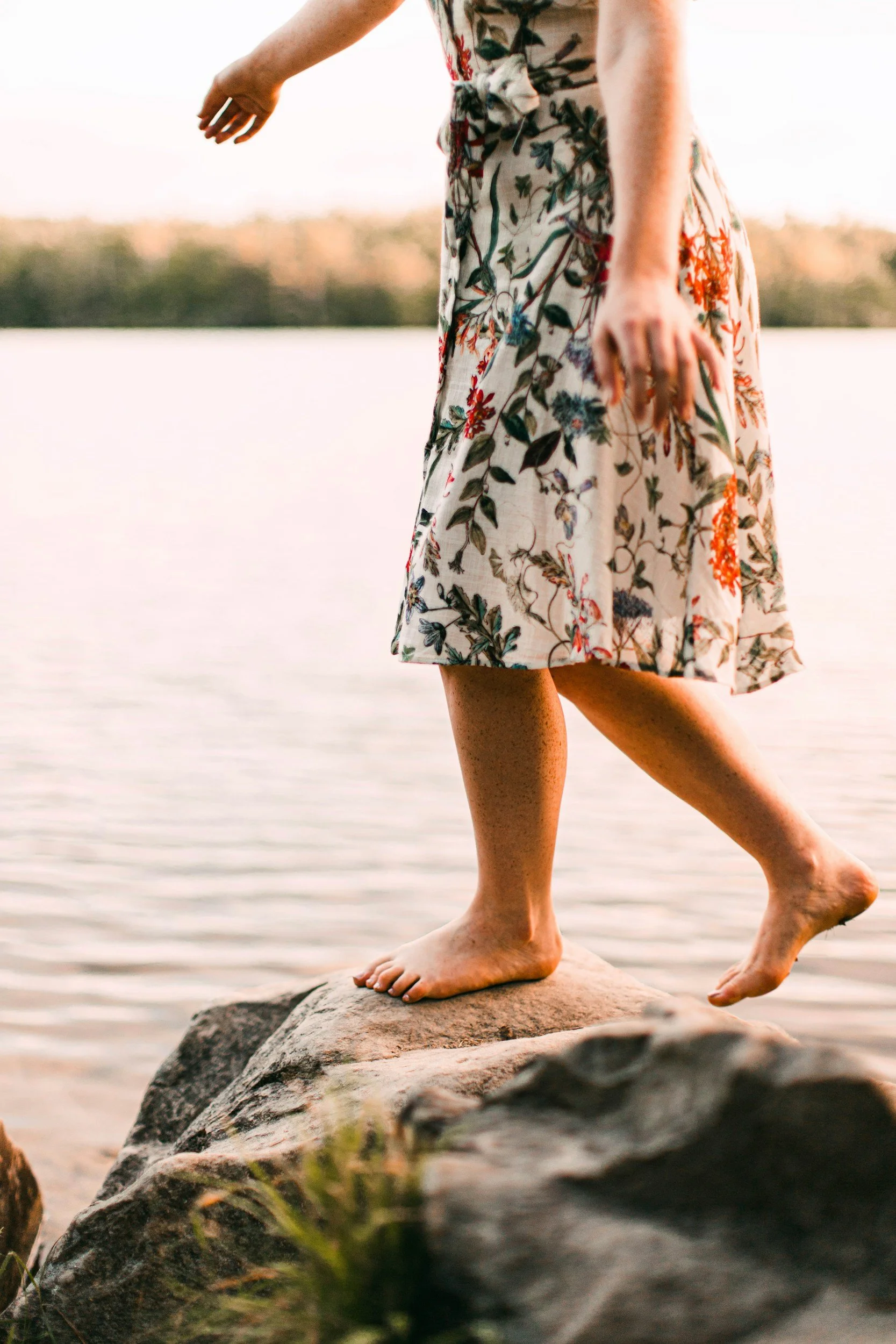 Person walking barefoot on rocks near a body of water during sunset, wearing a floral dress.