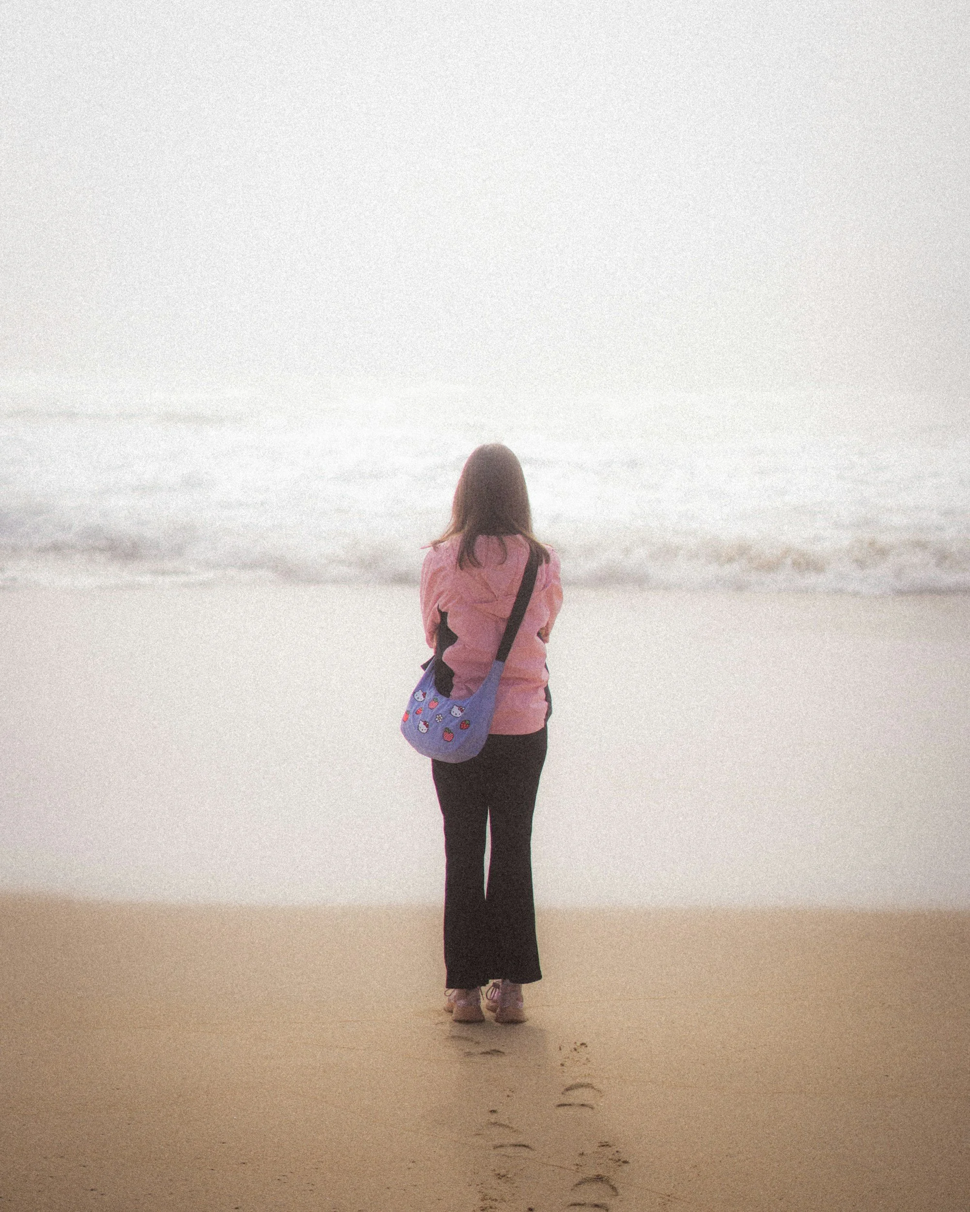 A woman standing on a sandy beach looking at the ocean, wearing a pink jacket, black pants, and a blue bag with red and white patterns.
