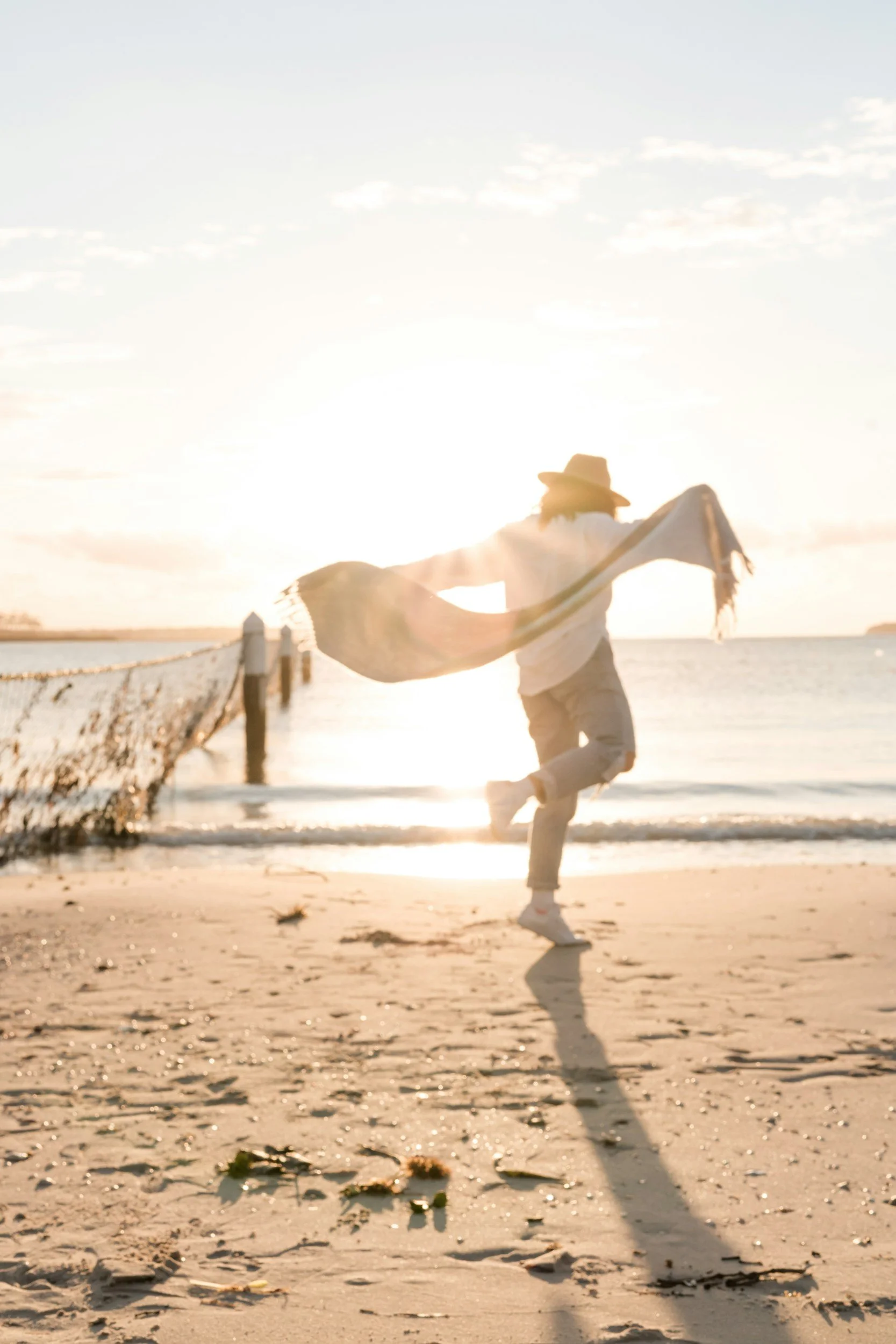 Person dancing on beach during sunset, wearing a hat and holding a cloth, with ocean and sky in the background.