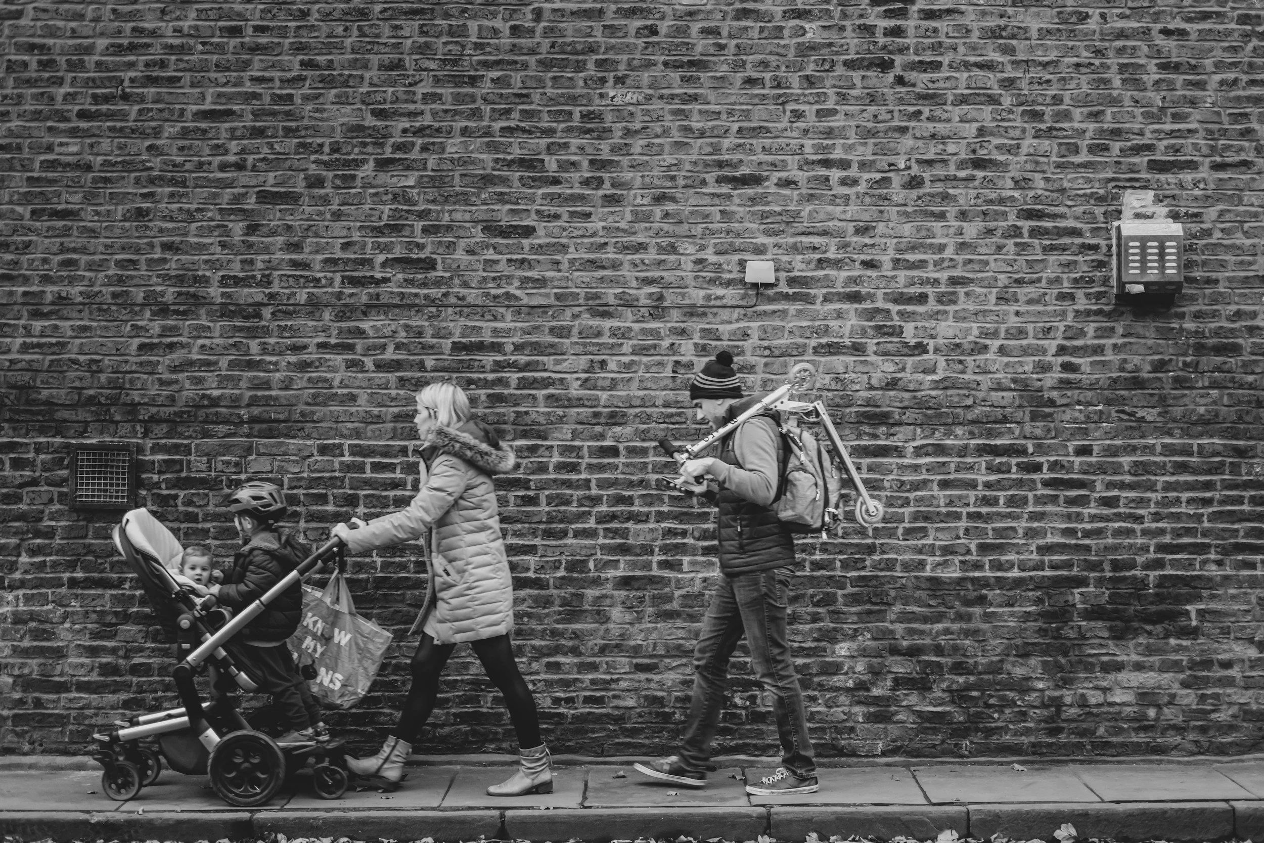 A black-and-white photograph showing several pedestrians walking along a sidewalk in front of a brick wall. The group includes a woman with a stroller, a man with a backpack carrying a bicycle, and a young girl with a striped beanie. The background features a large brick wall with some utility fixtures.