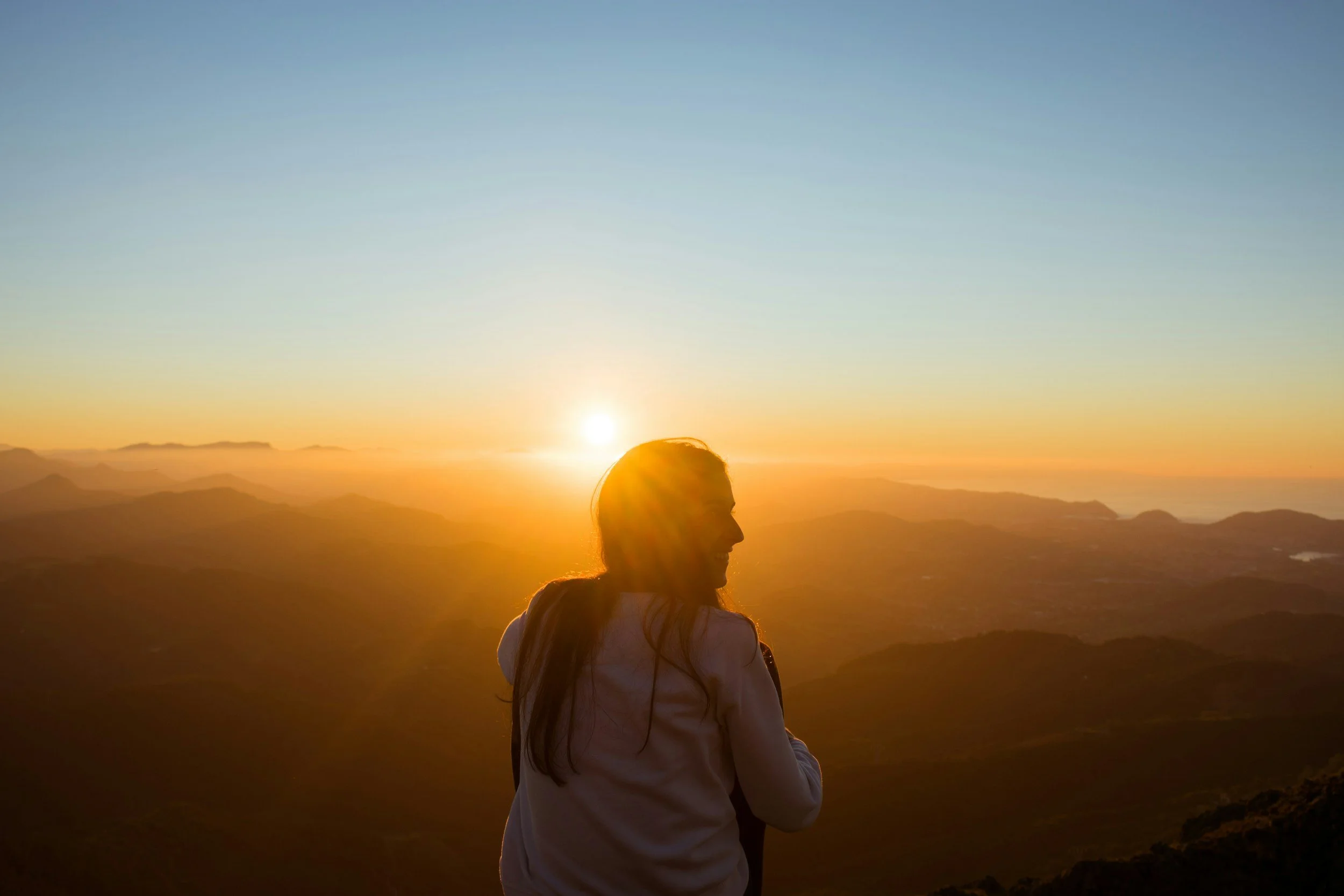 A woman with long hair sitting outdoors at sunset, with mountains in the background.