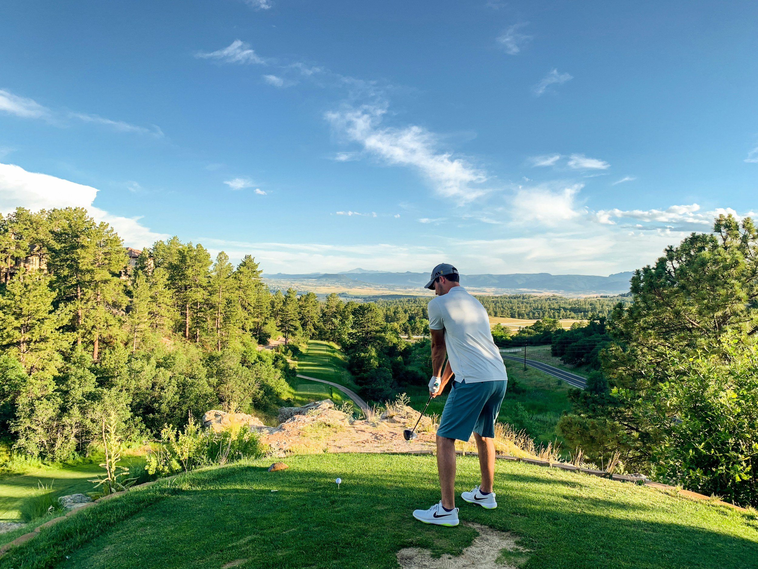 A man in a white t-shirt, blue shorts, and a cap preparing to hit a golf ball on a tee box surrounded by trees with a scenic view of a valley, mountains, and a blue sky with scattered clouds.