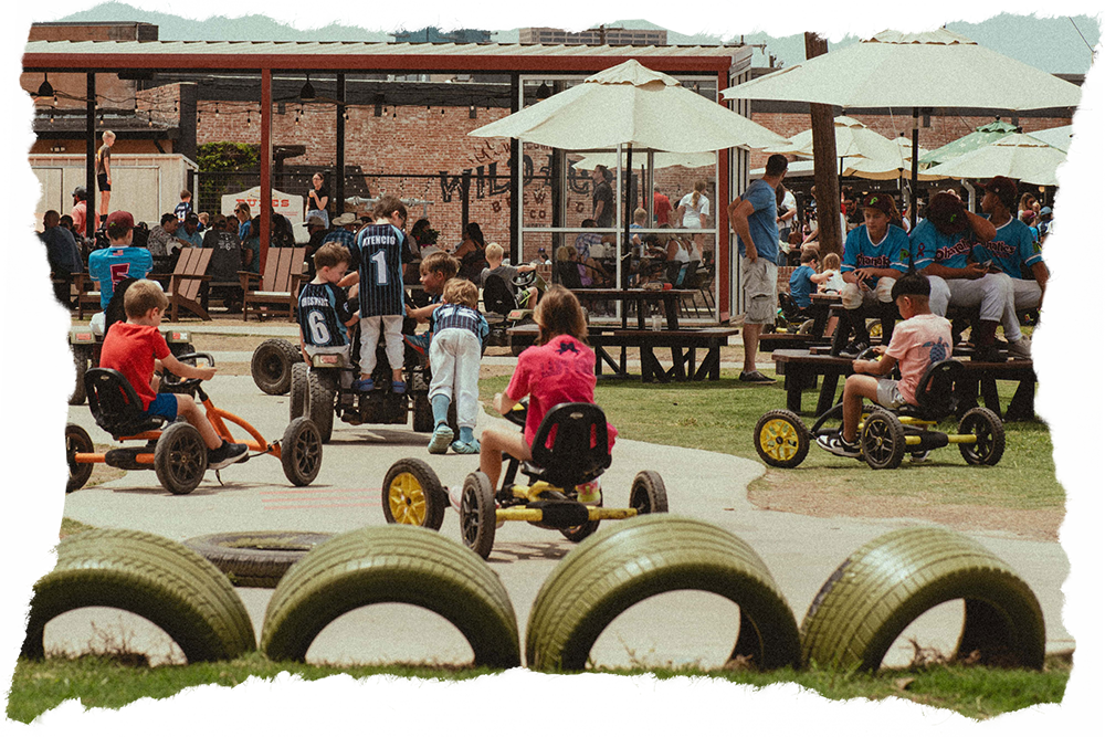 Children riding go-karts on a track, surrounded by people sitting at picnic tables under umbrellas in an outdoor area.