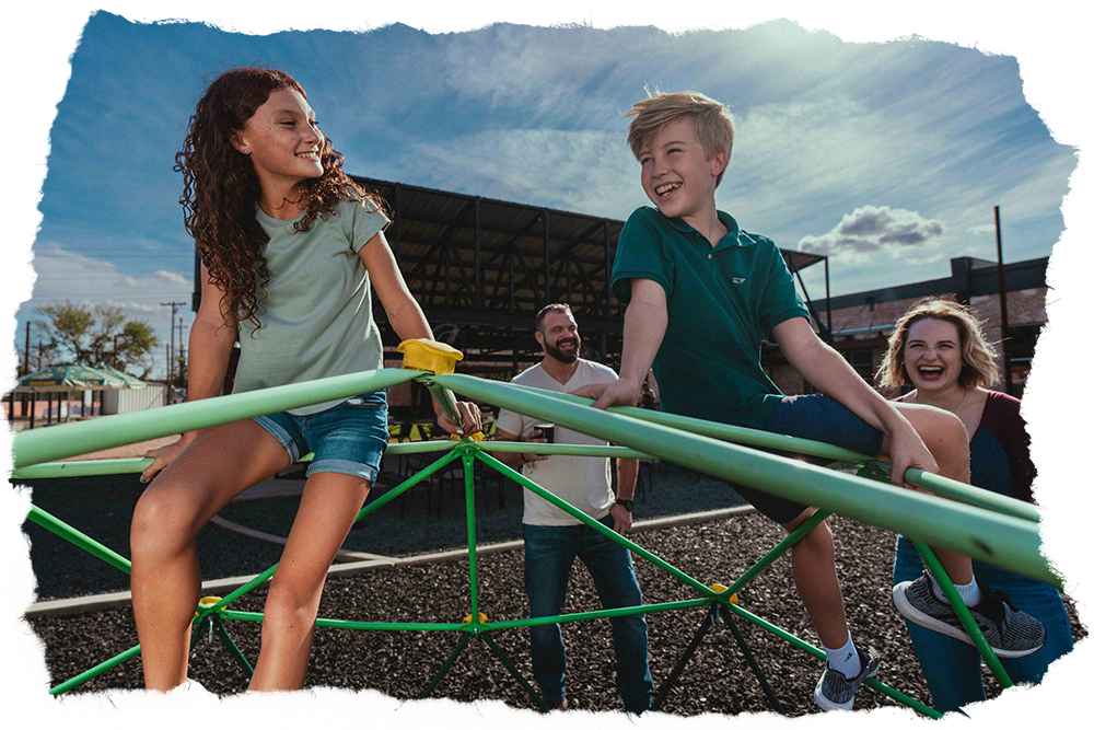 Two children climbing on playground equipment, with two adults nearby, enjoying a sunny day. The setting is an outdoor play area with clear skies.
