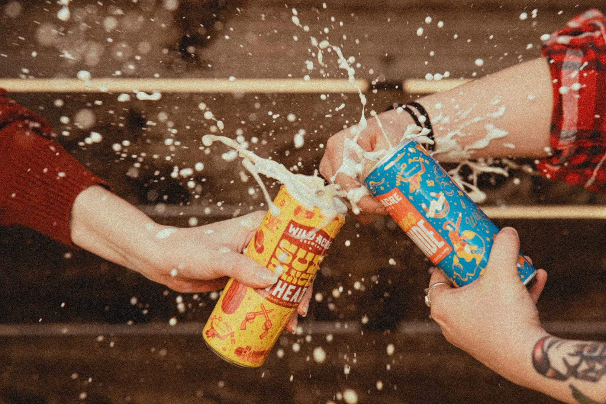 Two hands holding cans of beer with foam spraying out, one in a yellow can and the other in a blue can, against a wooden background.