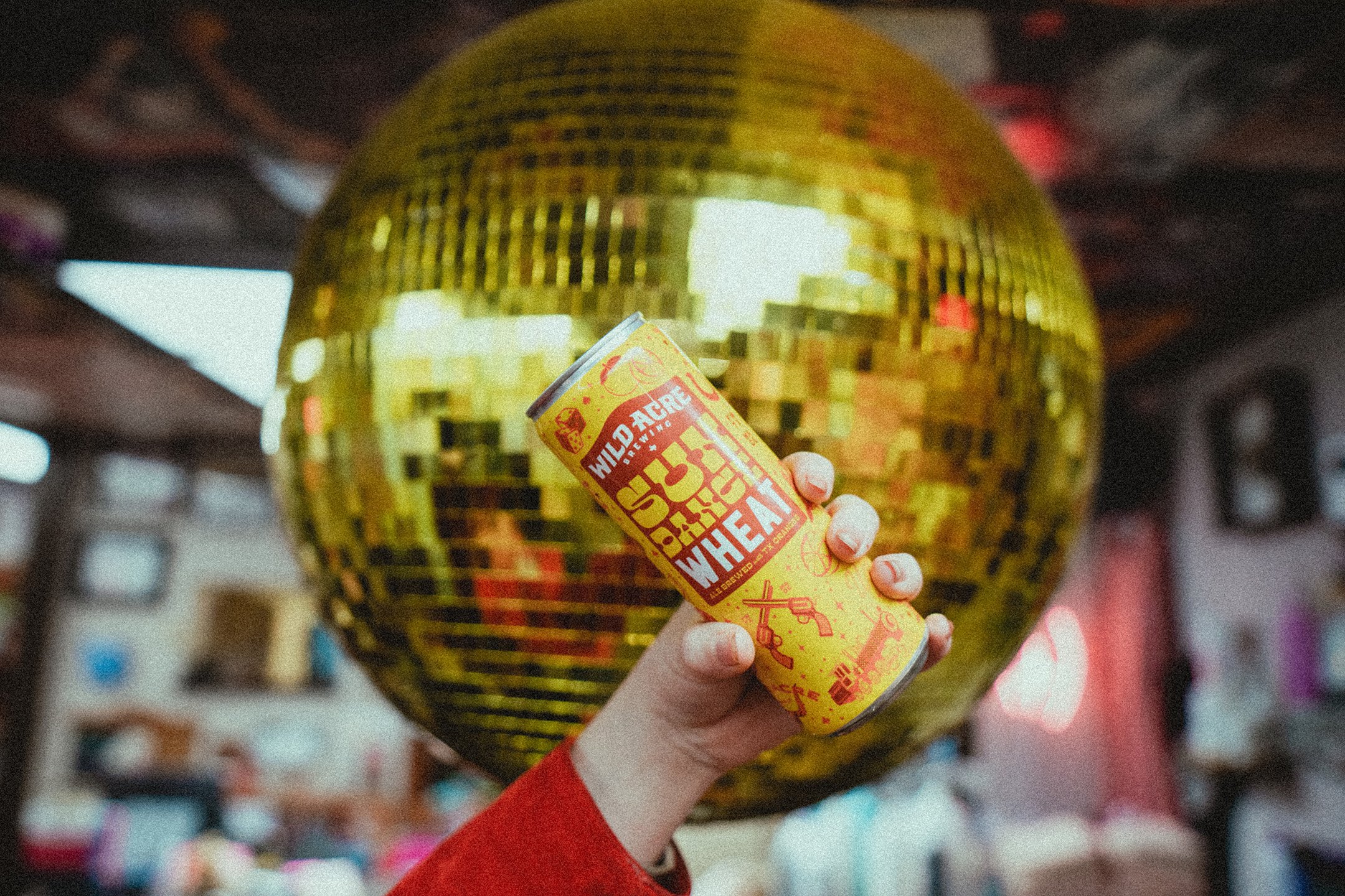 Person holding a Yellow Sulphur Wheat beer can in front of a large golden disco ball indoors.