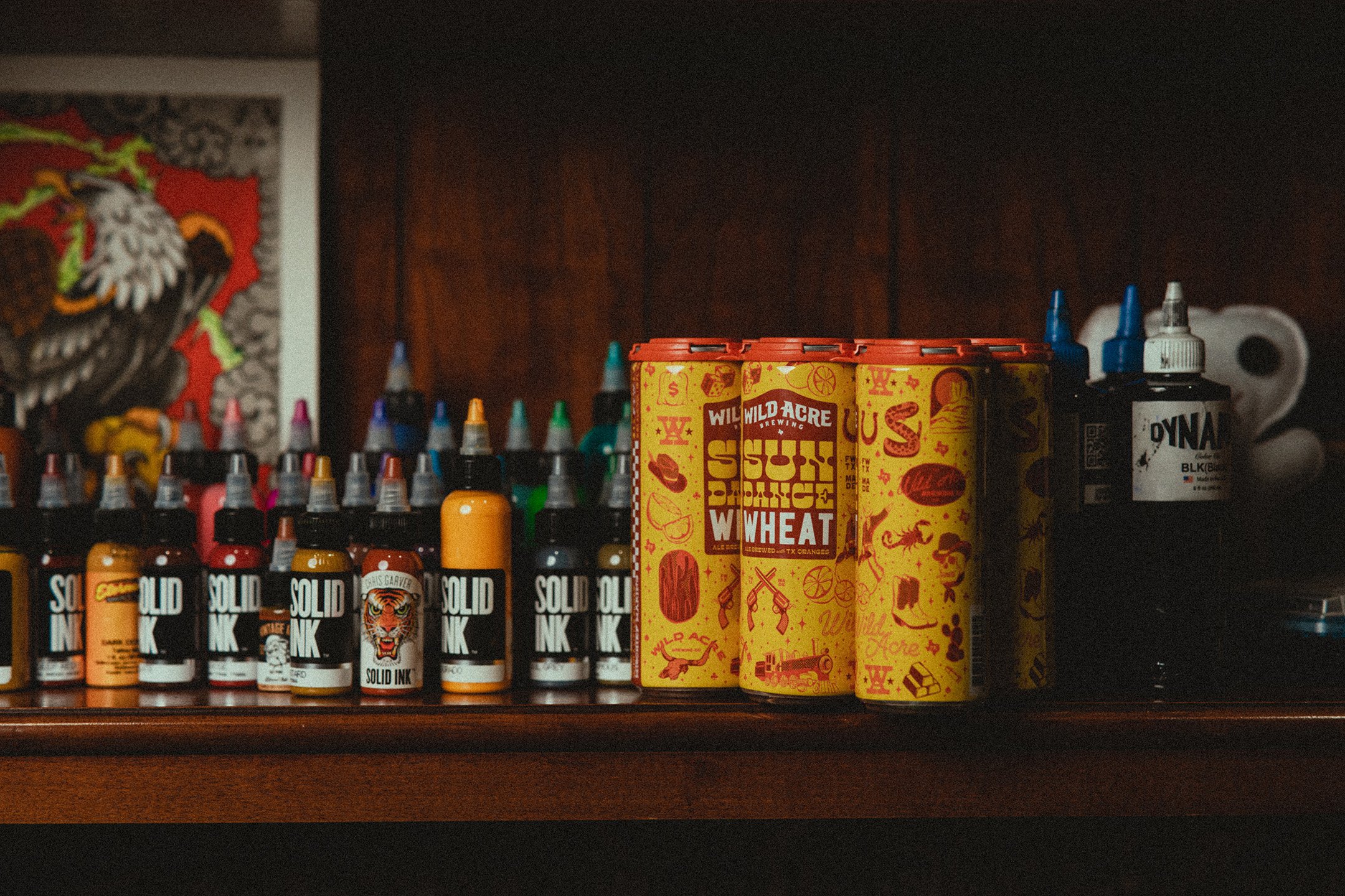 Assortment of colorful tattoo ink bottles and beer cans on a wooden shelf.
