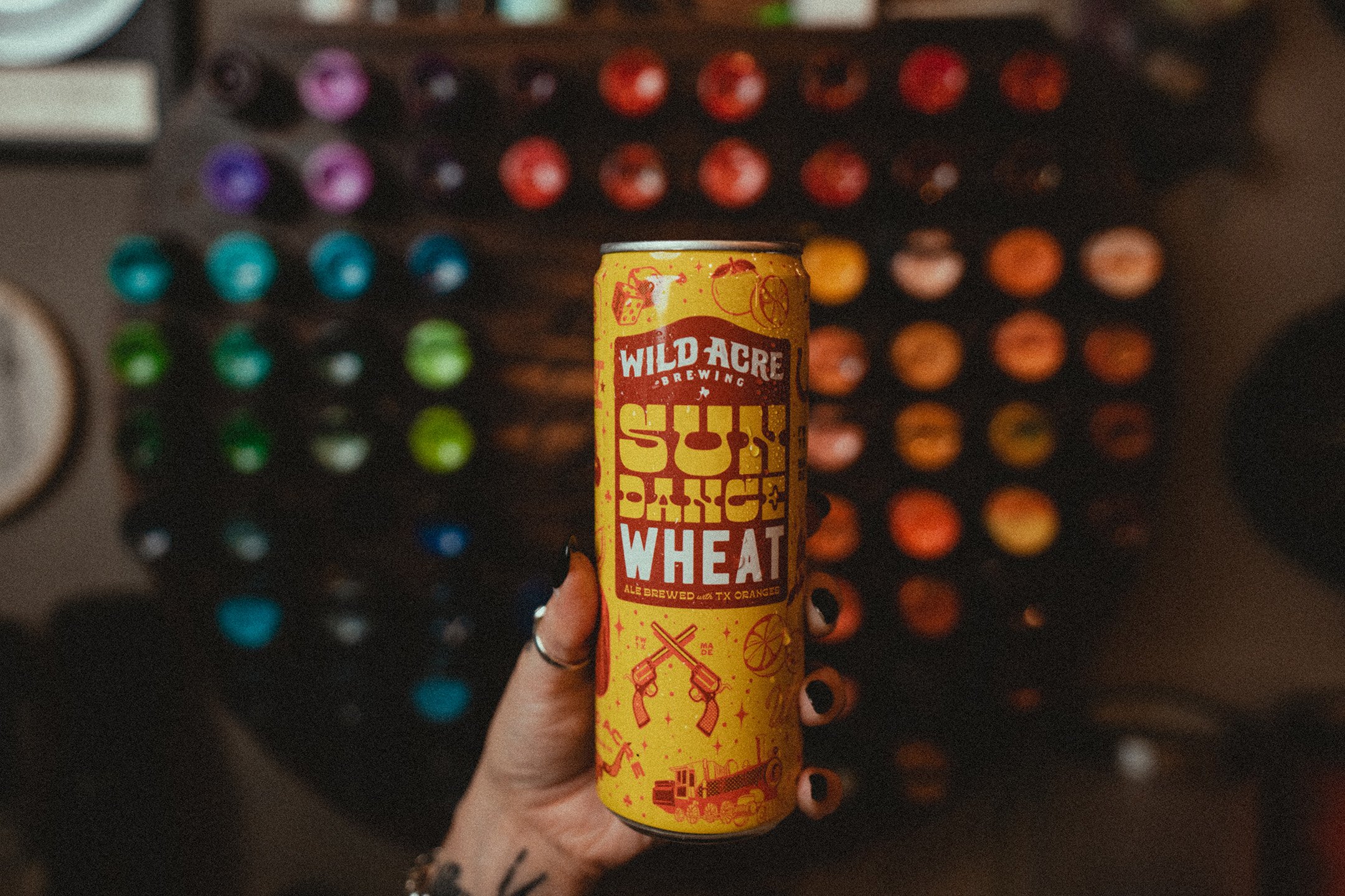 Hand holding a can of Wild Acre Brewing Sun Dance Wheat beer with colorful cans in the background.