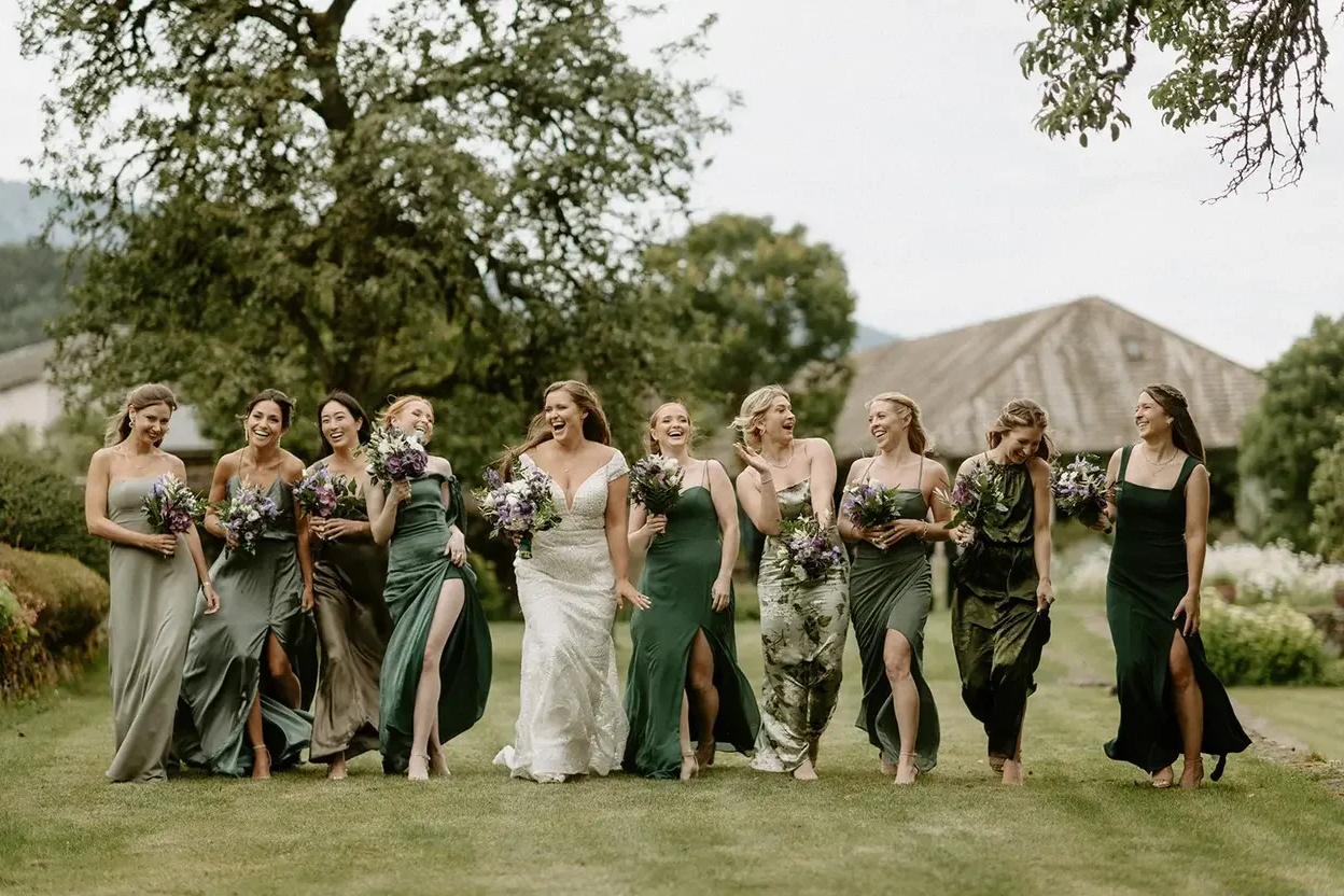 Group of women dressed in formal gowns walking on grassy field, smiling, and holding bouquets of flowers, outdoors with trees and a building in the background.