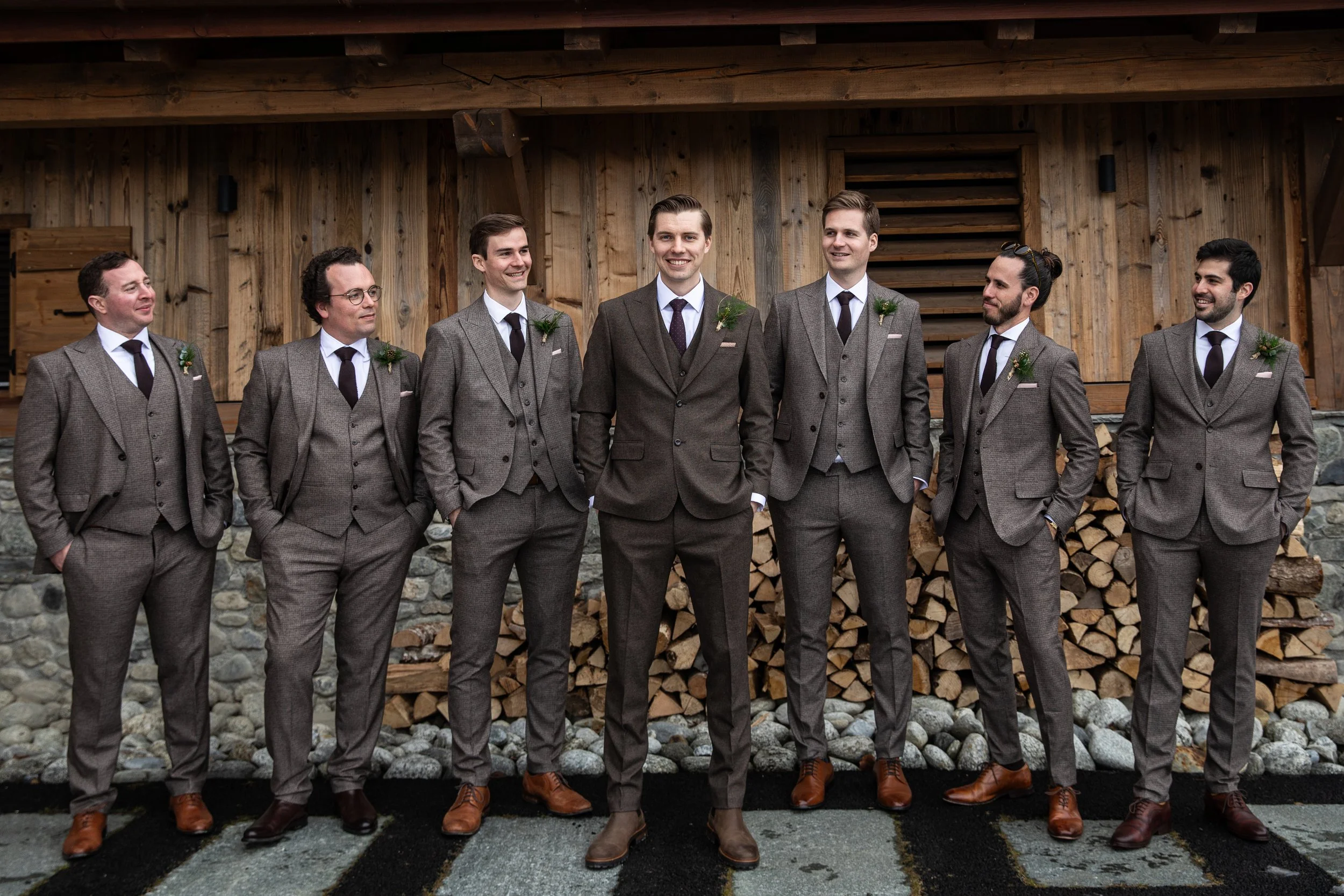 Group of seven men in gray suits standing outdoors in front of a wooden structure, smiling and looking at each other.