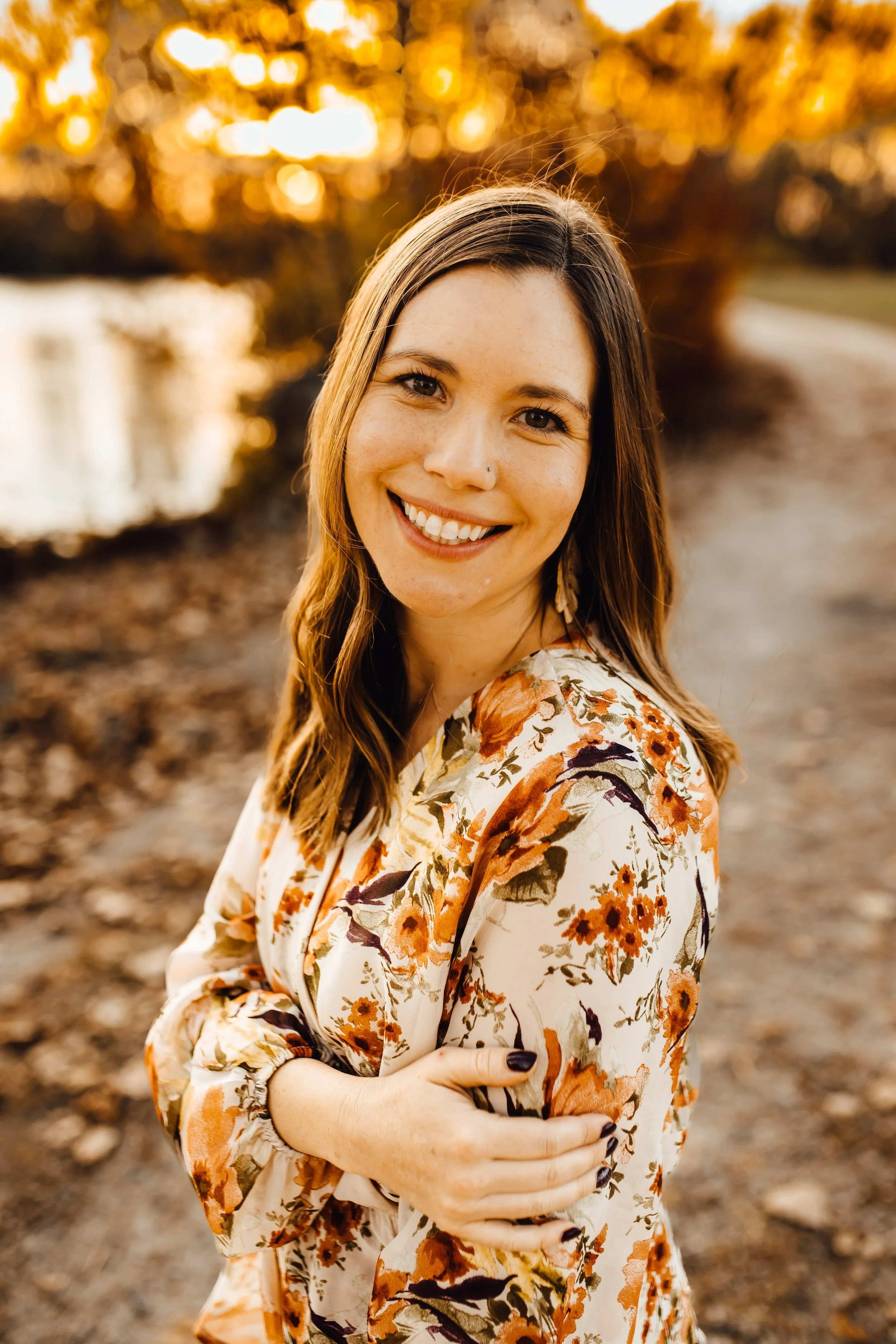 A woman with long brown hair smiling and wearing a floral dress, standing outdoors during sunset with a blurred background of trees and a path.