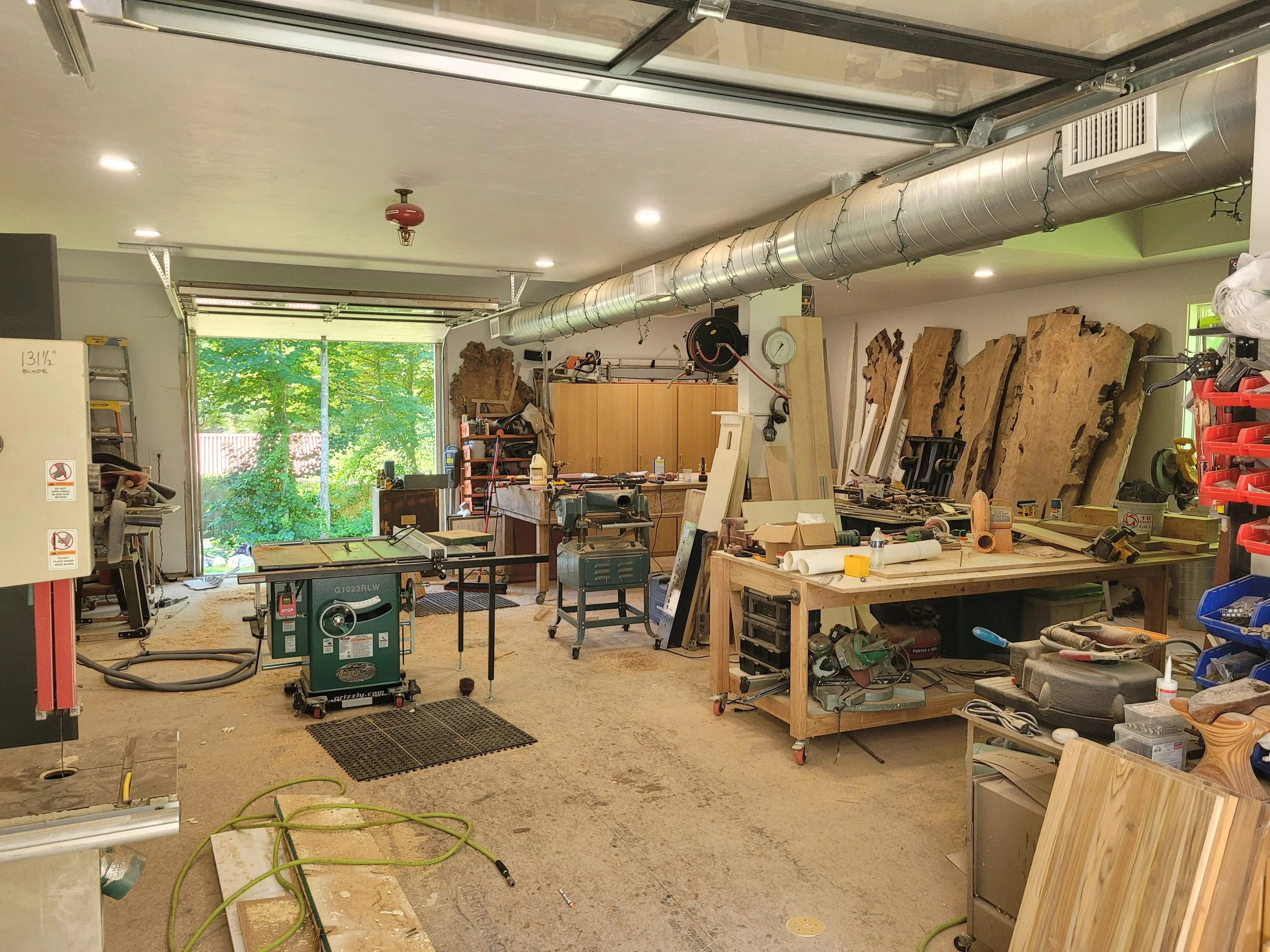 Woodworking workshop with various tools, large slabs of wood, and a work table, illuminated by overhead lights with a large open garage door showing green trees outside.