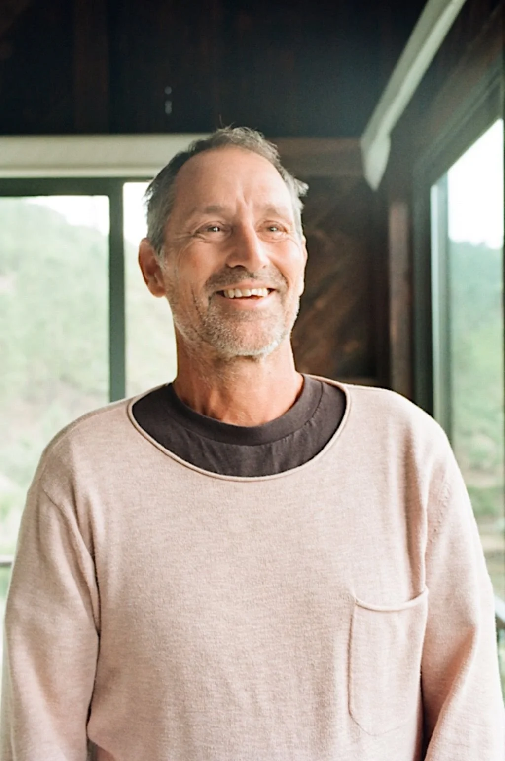 Man smiling indoors near a window, wearing a light pink sweater over a dark shirt.