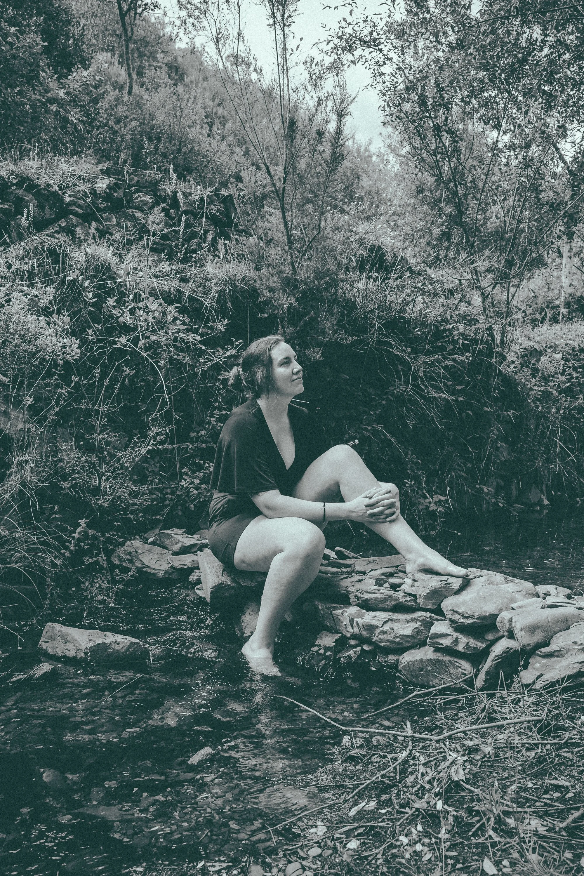 Woman sitting on rocks by a stream in a lush forest area.