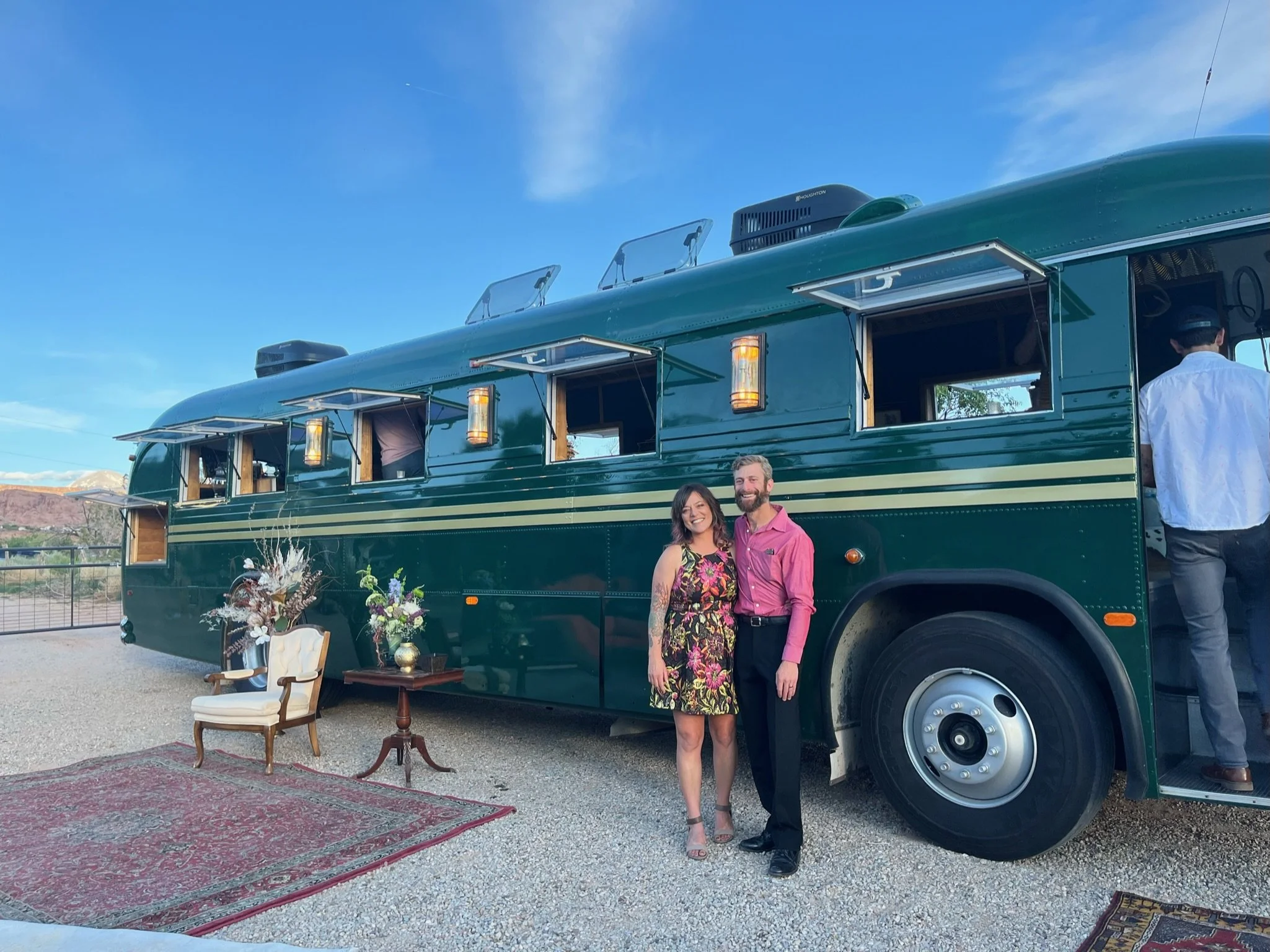 Couple standing in front of a vintage green bus with open windows, tables, chairs, and decor outside on a gravel area, with a mountain scenic background under a bright blue sky.